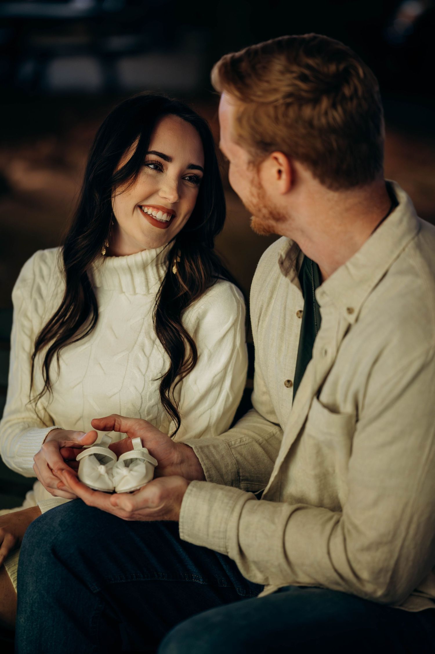Smiling woman and man holding tiny white baby booties together during a Christmas pregnancy announcement photo.