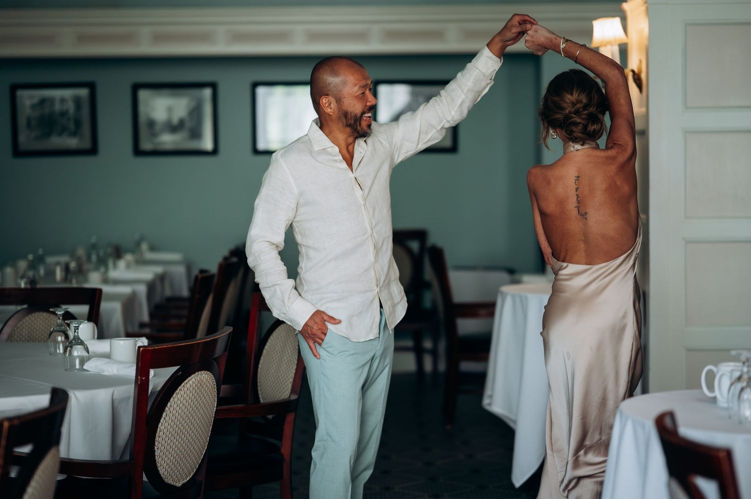 A couple dances elegantly in a restaurant, the bride in a backless gown as the groom twirls her gracefully.