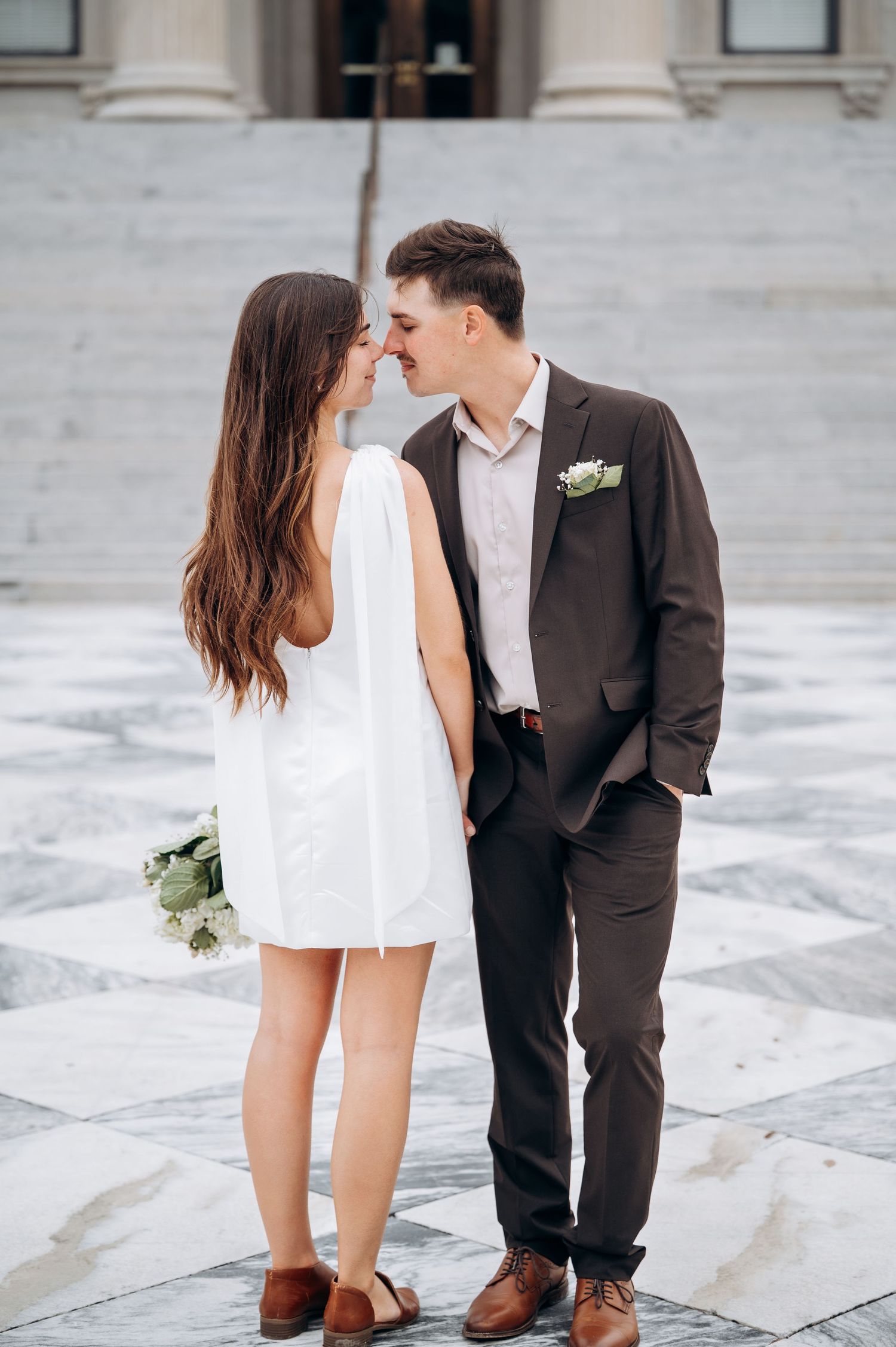 Bride and groom lean in for a kiss on marble steps outside a grand building on their wedding day.