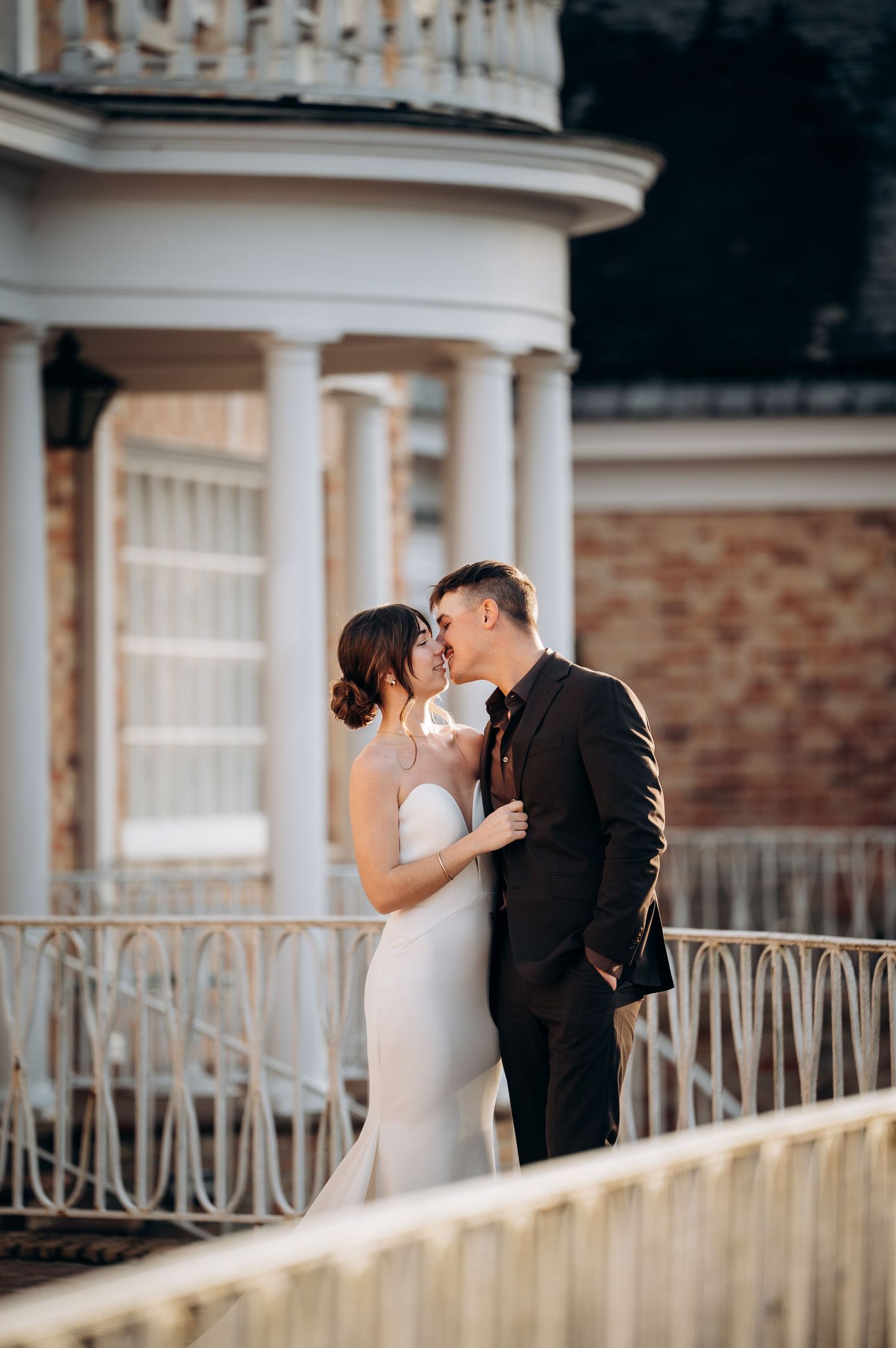 Bride and groom share a romantic kiss on a balcony with classical columns of an estate behind them.