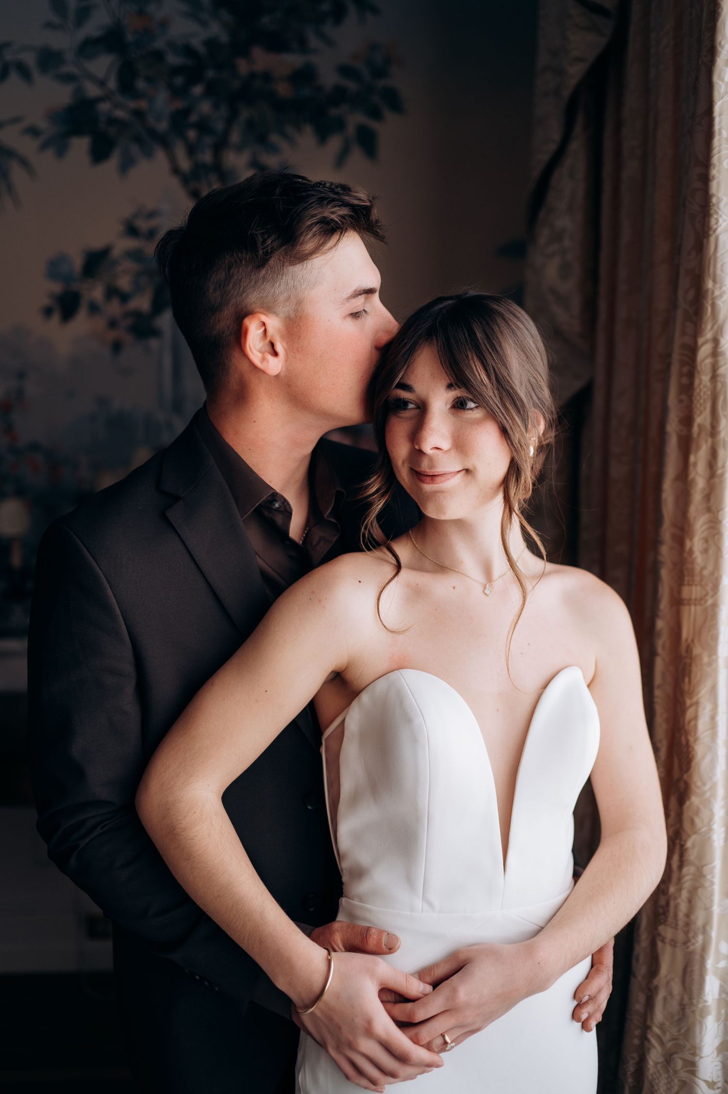 Elegant couple in formal attire pose against vintage wallpaper in soft lighting.