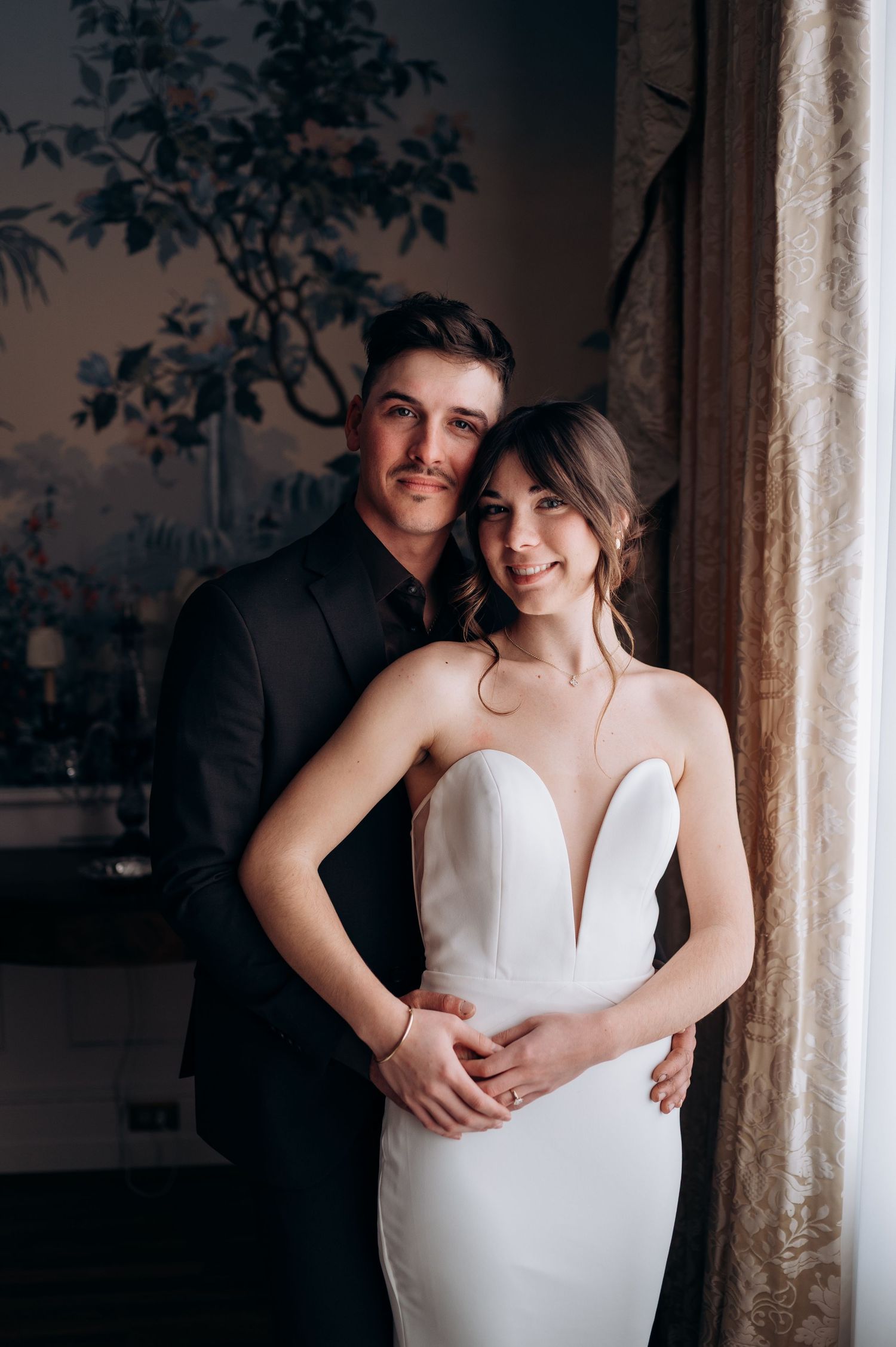 A tender moment captured between a couple in formal attire against a scenic wallpapered background.