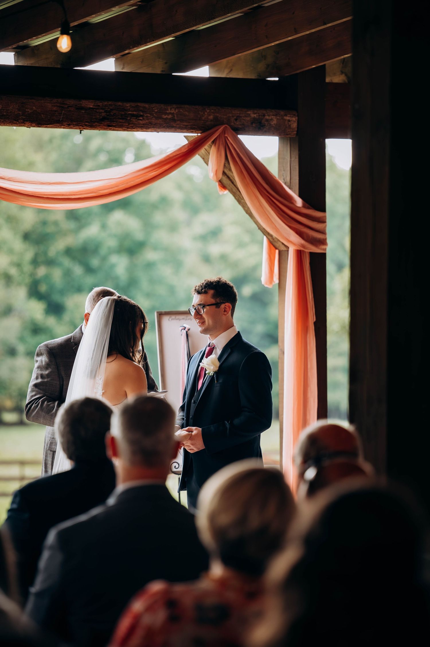 Wedding ceremony in progress under wooden beams with orange draping and natural lighting.