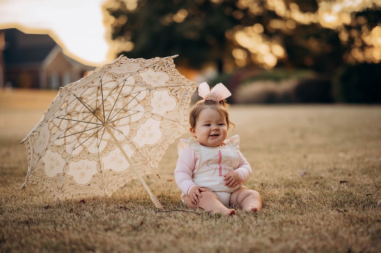 A baby in white sits on grass next to a delicate lace parasol during golden hour.