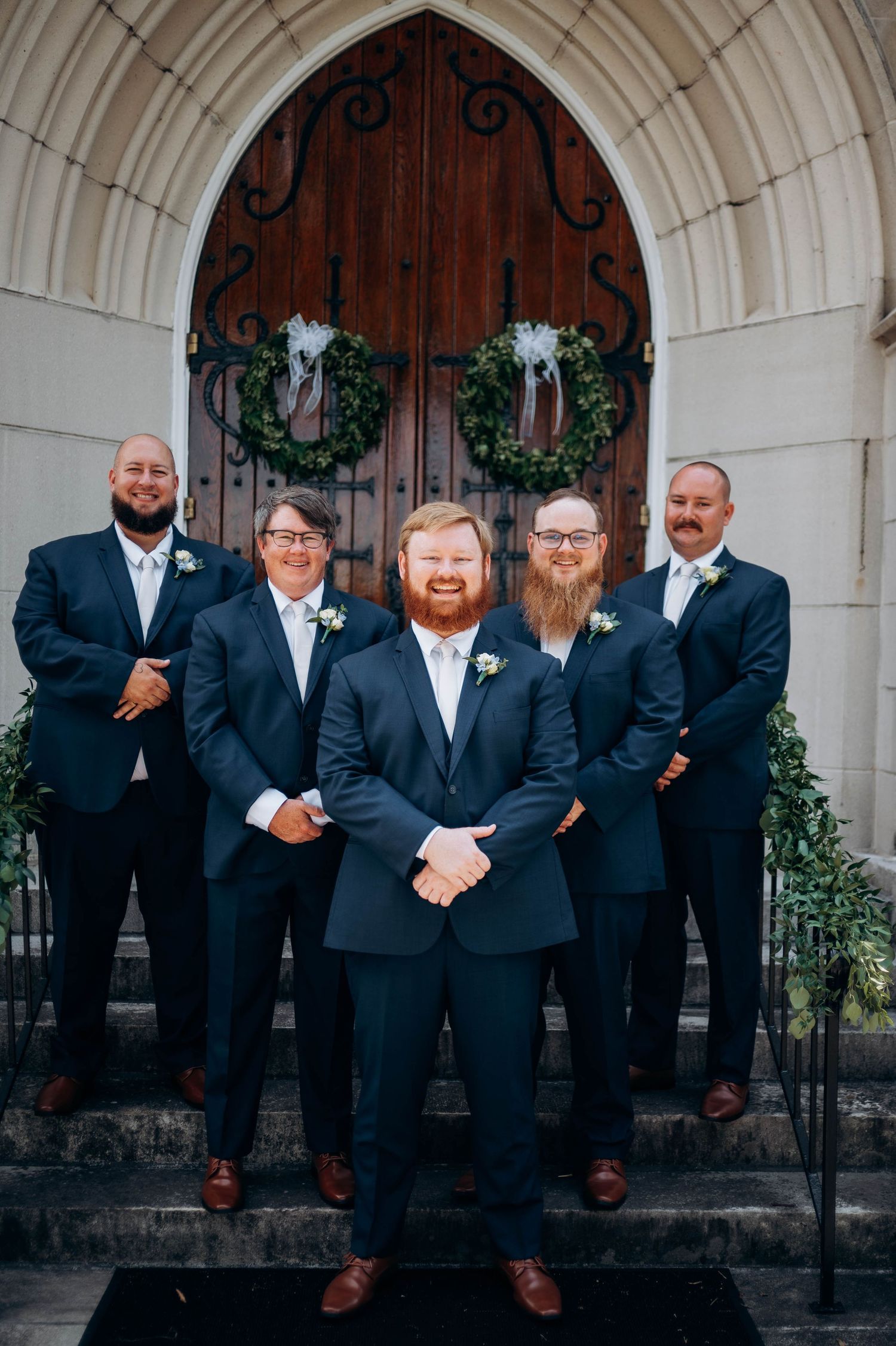 Groomsmen in dark suits pose on church steps decorated with wreaths.