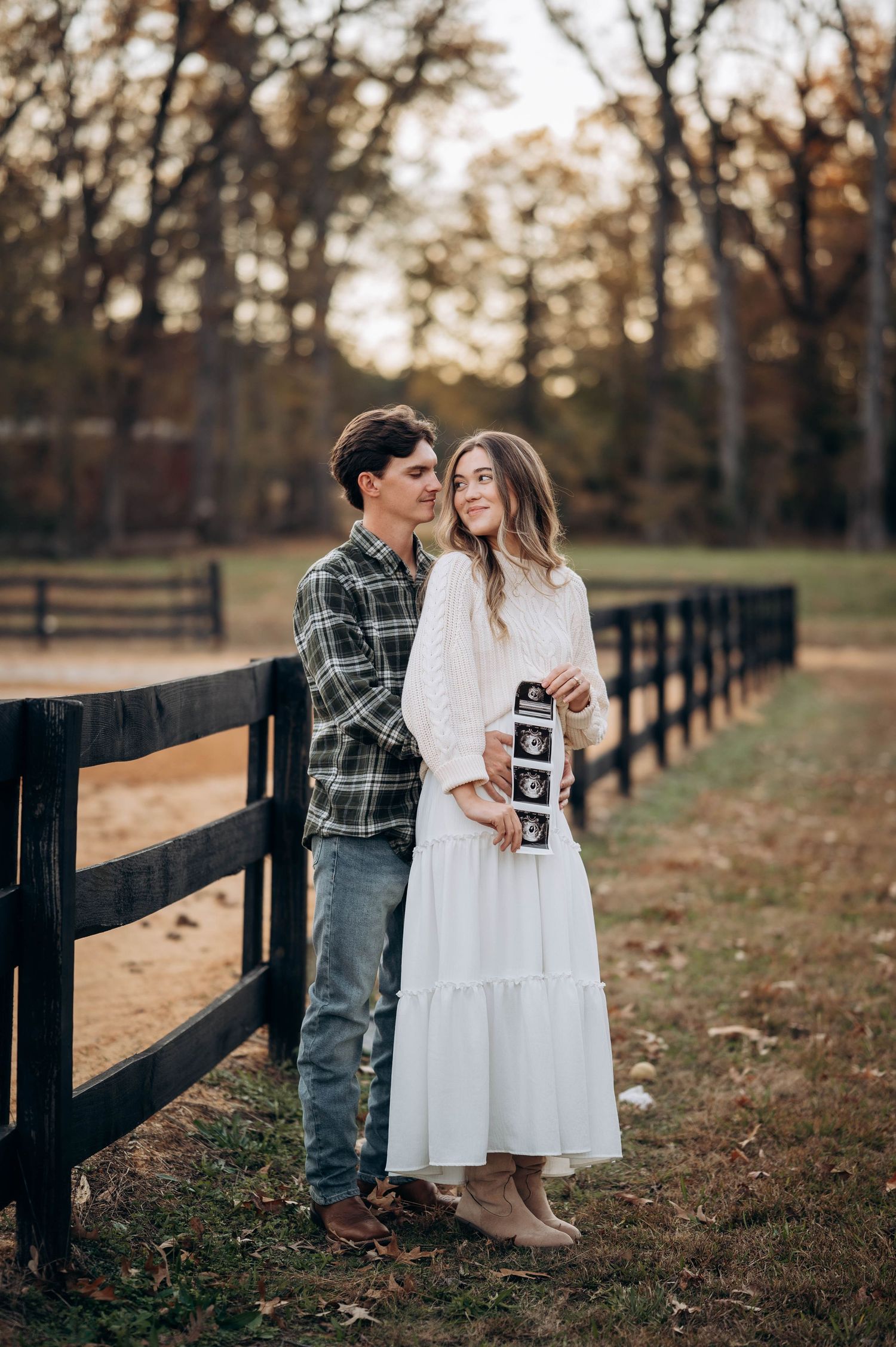 A couple poses along a wooden fence holding ultrasound photos during a fall pregnancy announcement.