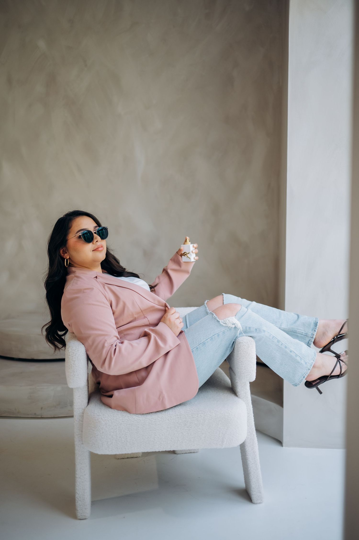 Stylish outfit featuring pink blazer, ripped jeans and sunglasses while lounging in white chair.