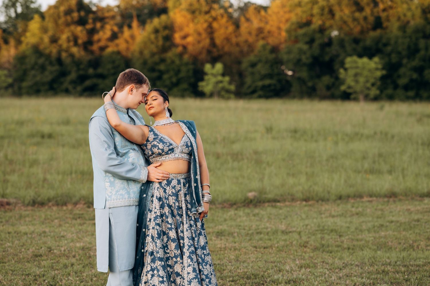 Couple embracing in a green field, wearing elegant blue wedding attire, with autumn trees in the background.