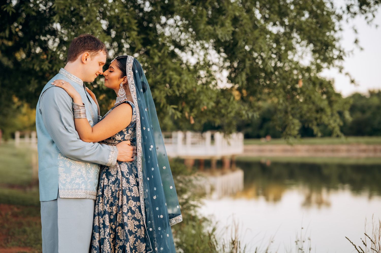 Couple in traditional South Asian wedding attire embracing near a serene lake surrounded by lush green trees.