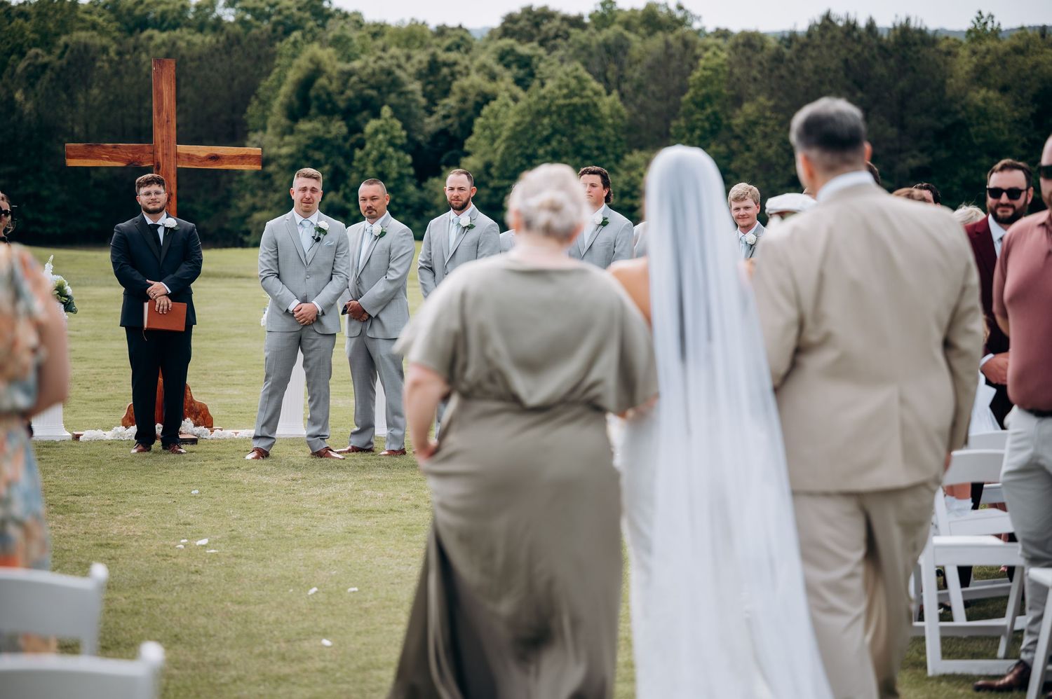 Bride walks down outdoor aisle with groomsmen in grey suits awaiting at cross altar on green lawn.