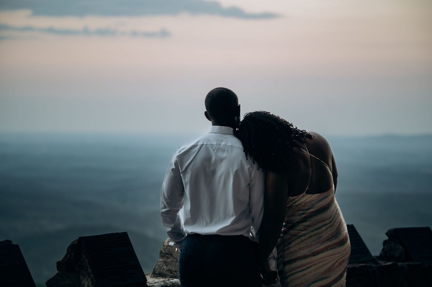Romantic couple embracing at scenic overlook during twilight, man in white shirt and woman leaning on his shoulder.