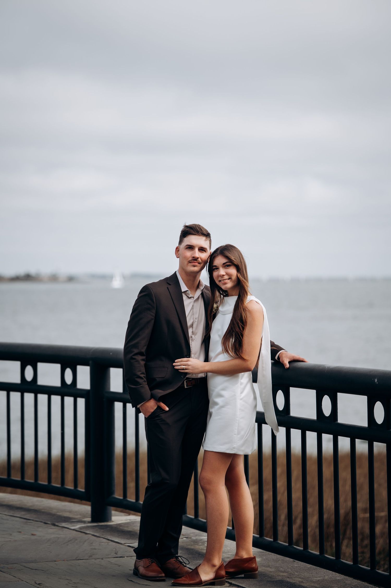 Elegant couple poses by waterfront railing on cloudy day, bride in white mini dress and groom in suit.