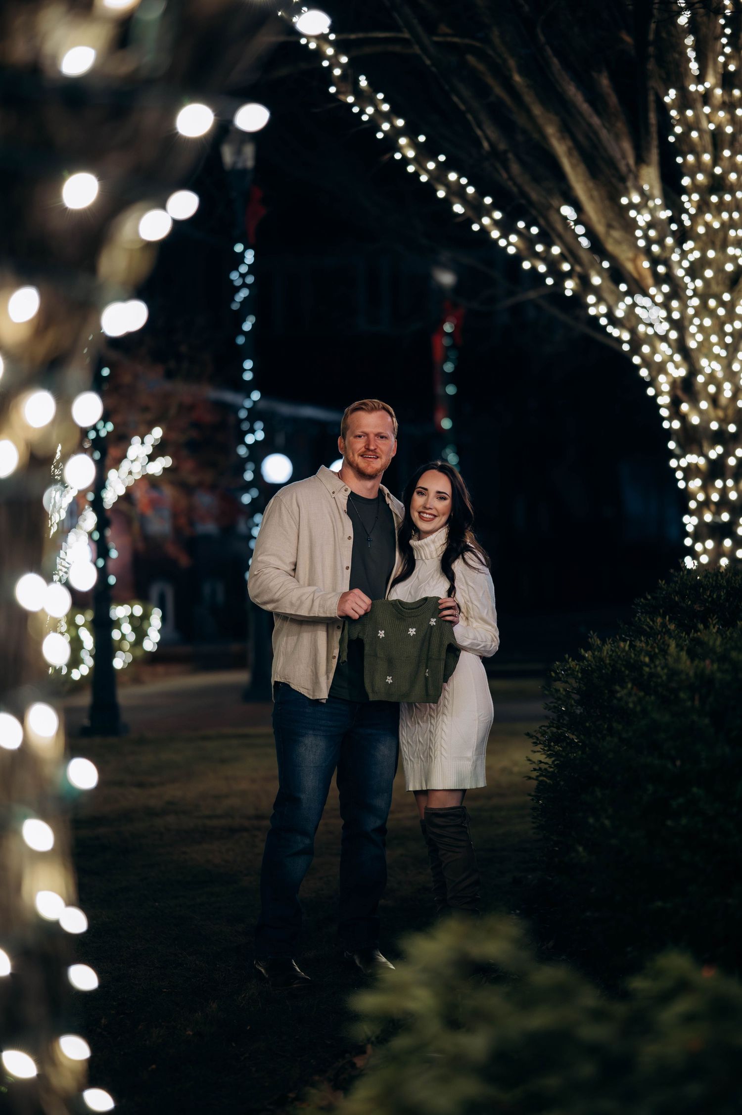 Couple smiling together under twinkling Christmas lights on trees at night, holding a green bag.