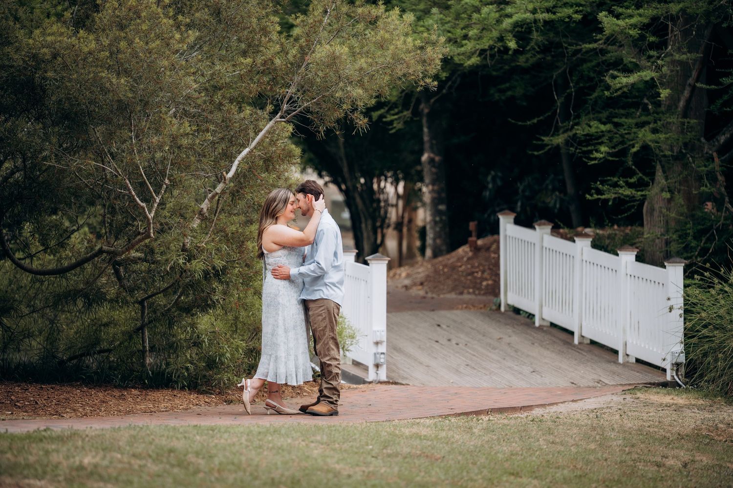 Couple embracing and kissing near white fence gate surrounded by lush green trees outdoors.