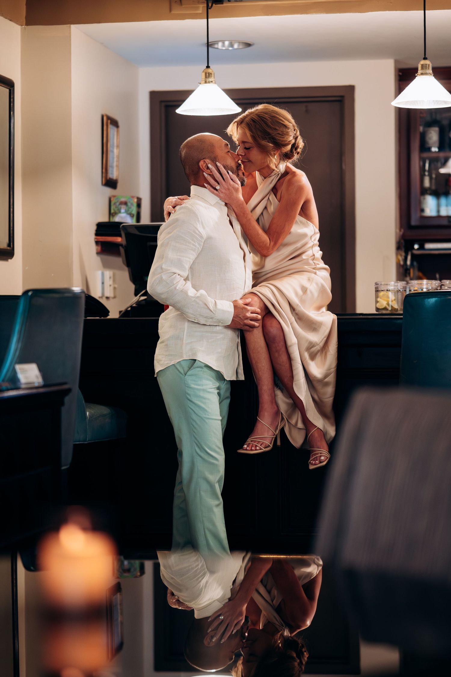 Romantic couple kissing at a dimly lit bar, man in white suit and woman in satin gown seated on counter.