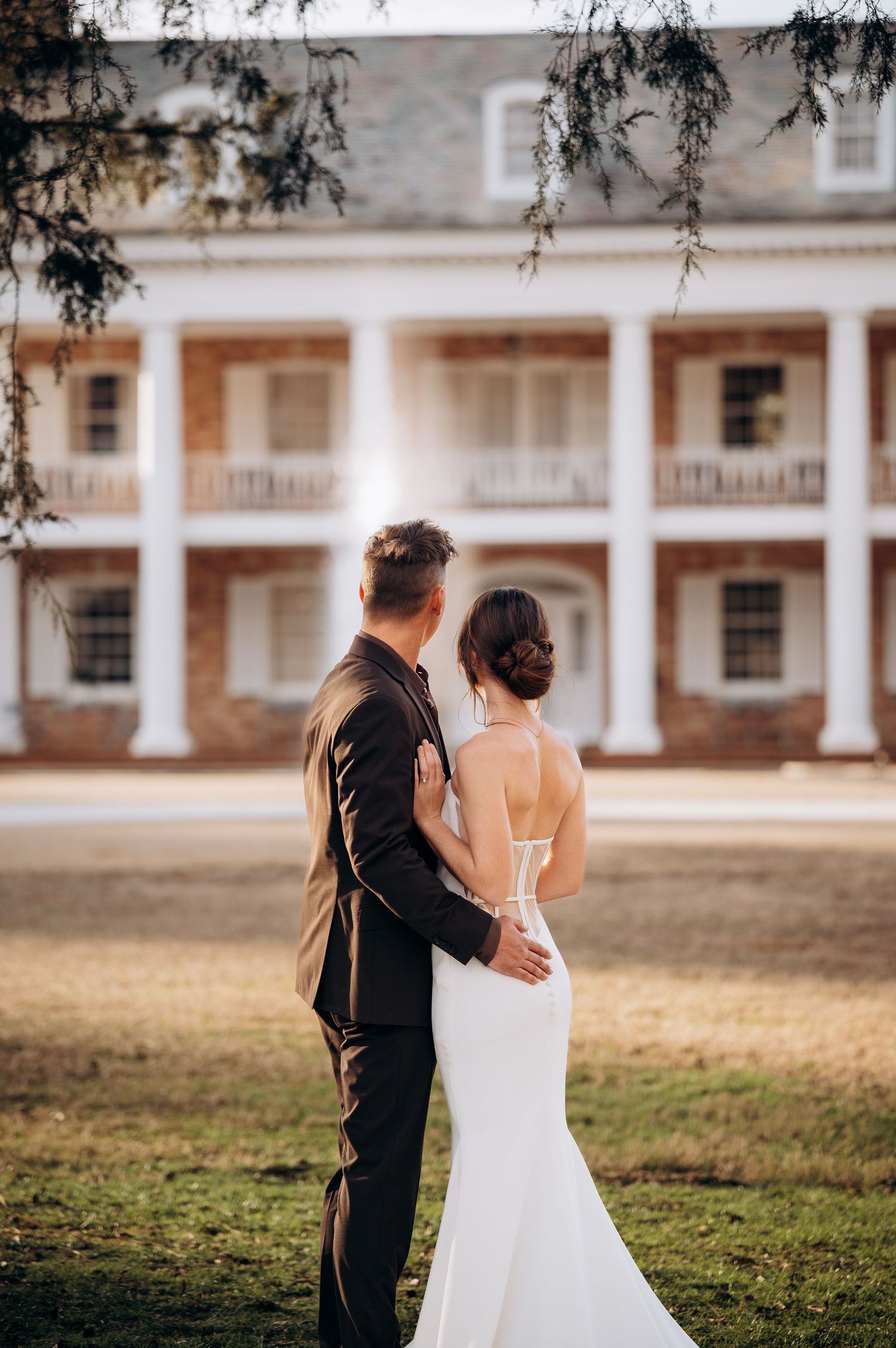 Bride and groom stand together gazing at an elegant Southern plantation mansion with white columns.