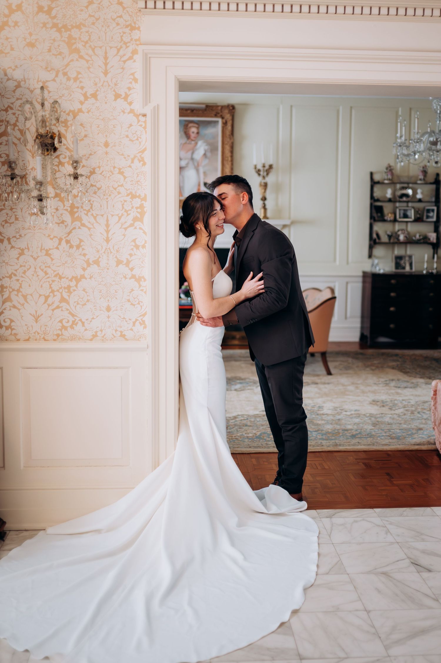 Bride and groom embracing in doorway of elegant room with ornate wallpaper and classic portrait painting.