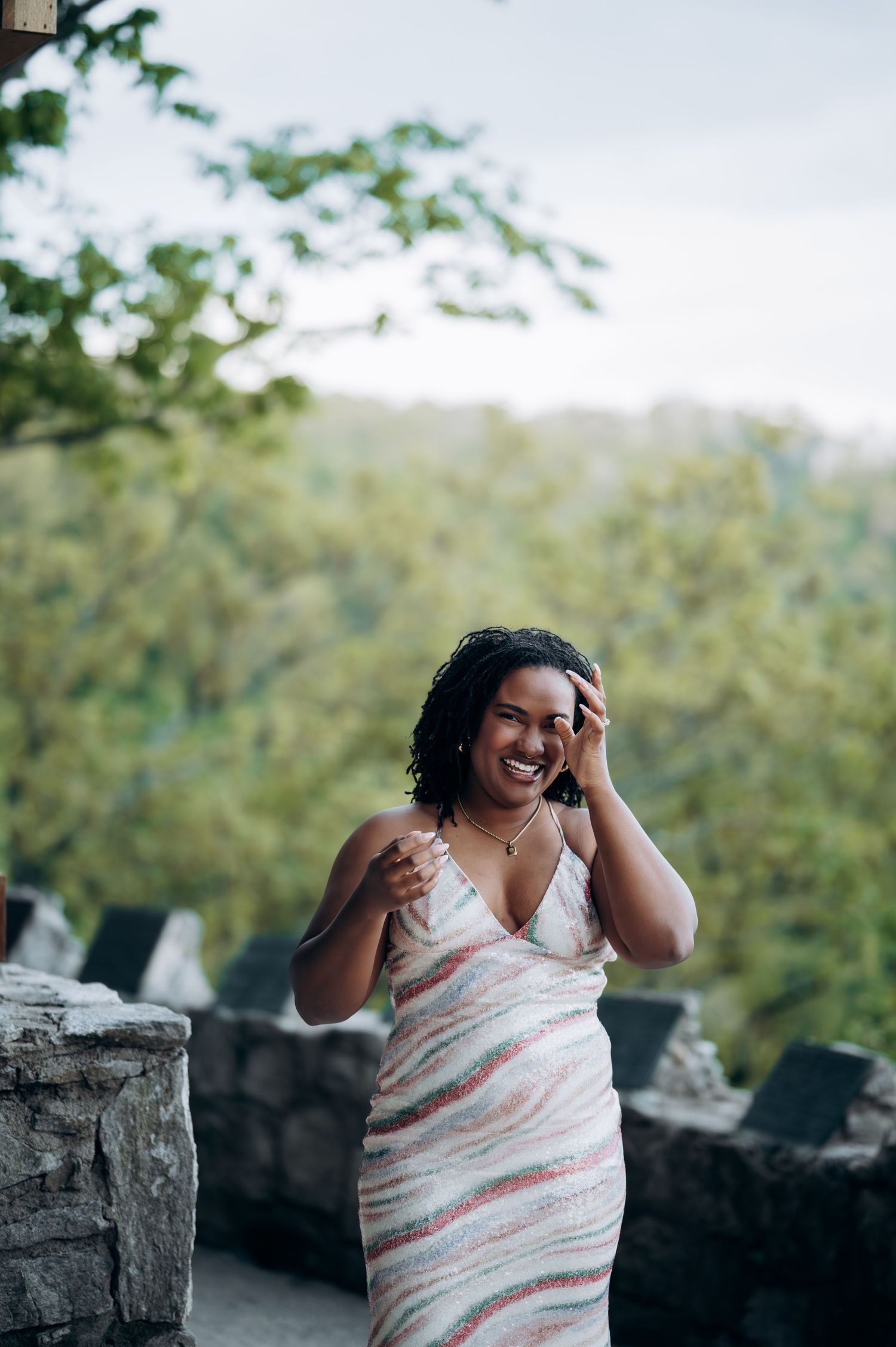 A smiling Black woman in a striped dress laughs outdoors near stone walls with lush green trees in the background.