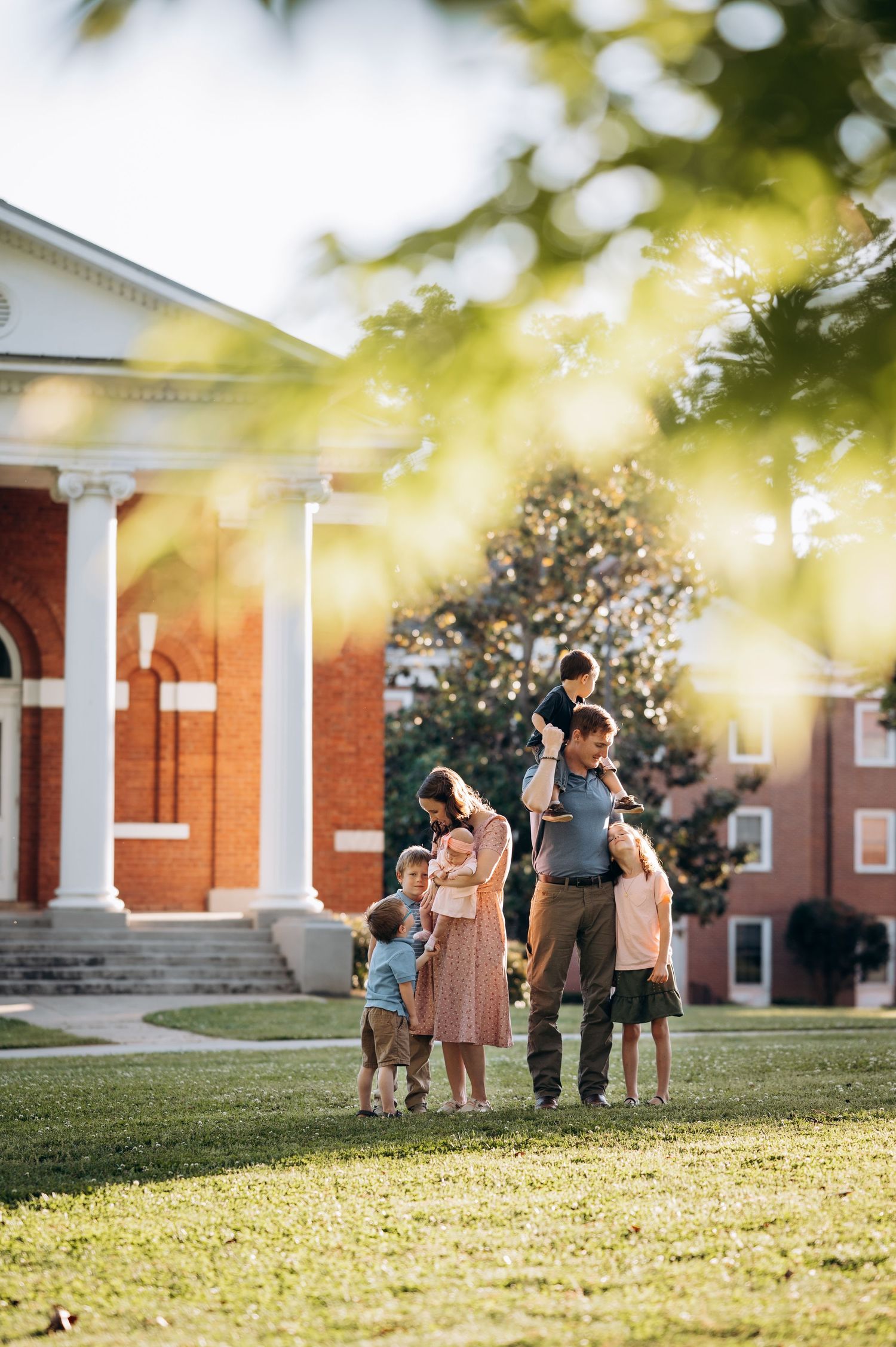 Family with young children gathered on green lawn outside a historic red brick building with white columns on a sunny day.