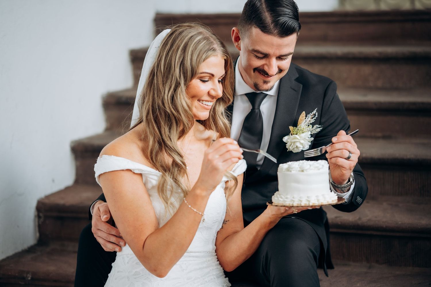 Bride and groom cutting a small white wedding cake together while sitting on wooden stairs.