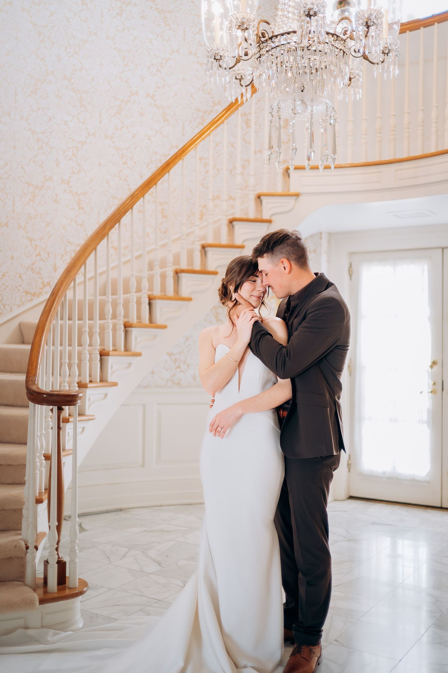Couple shares romantic moment under crystal chandelier by curved staircase.
