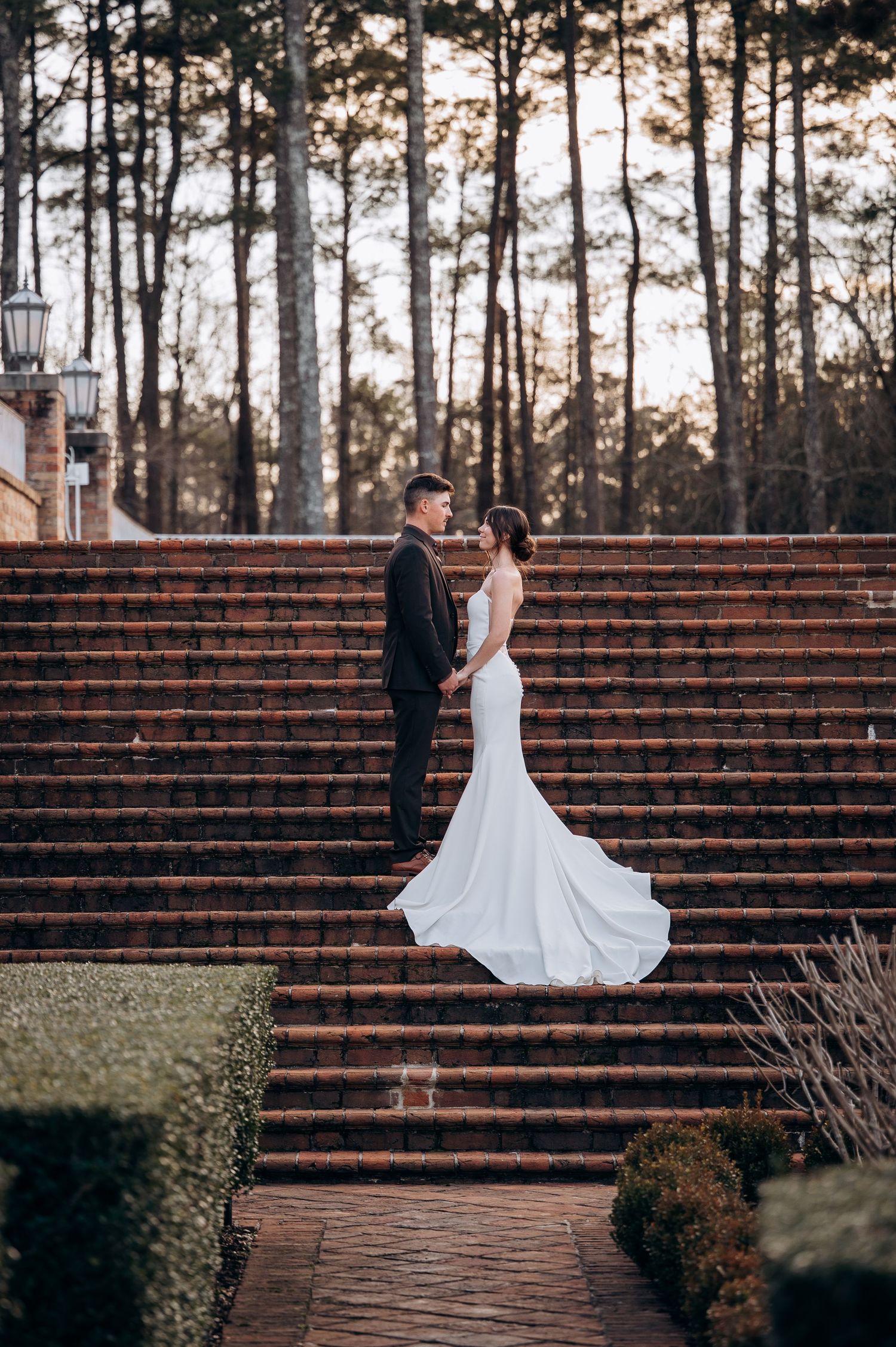 Wedding couple poses on grand brick steps with pine forest backdrop at sunset.