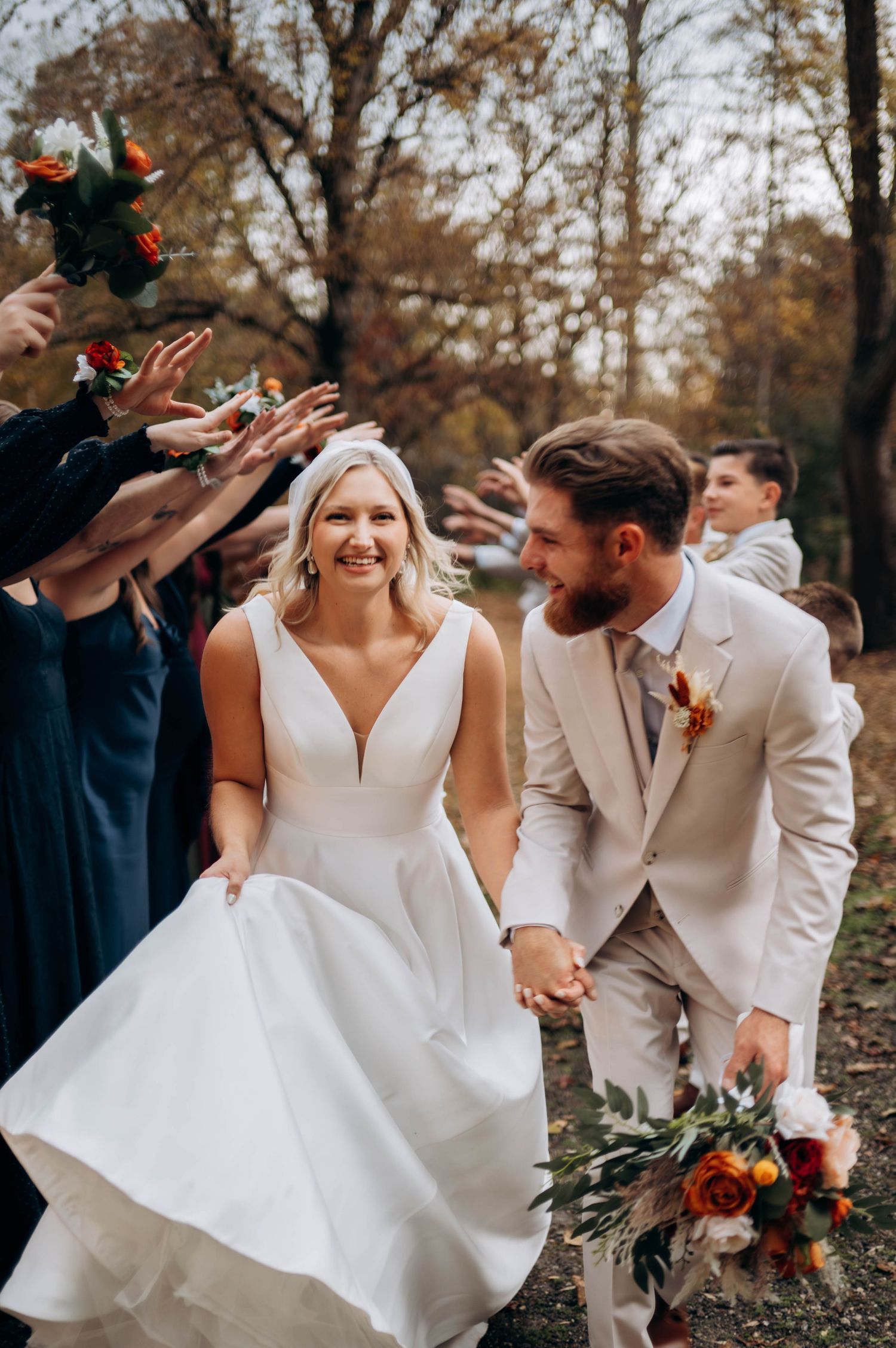 Joyful wedding couple laughs together during outdoor autumn photoshoot in matching white and beige attire.