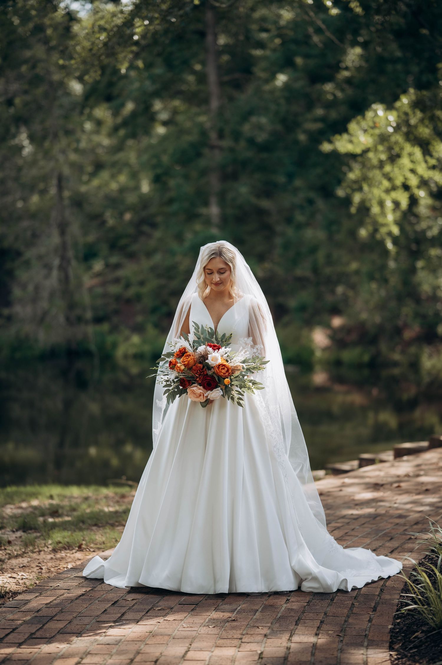Bride in flowing white gown sits on brick path holding fall-colored bouquet.