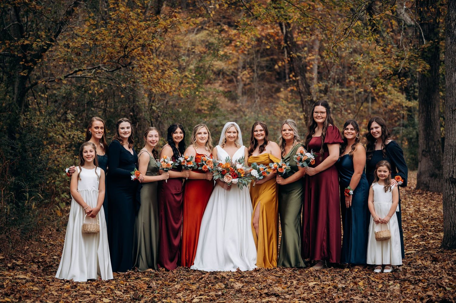 A wedding party in fall-colored dresses poses outdoors among autumn foliage.