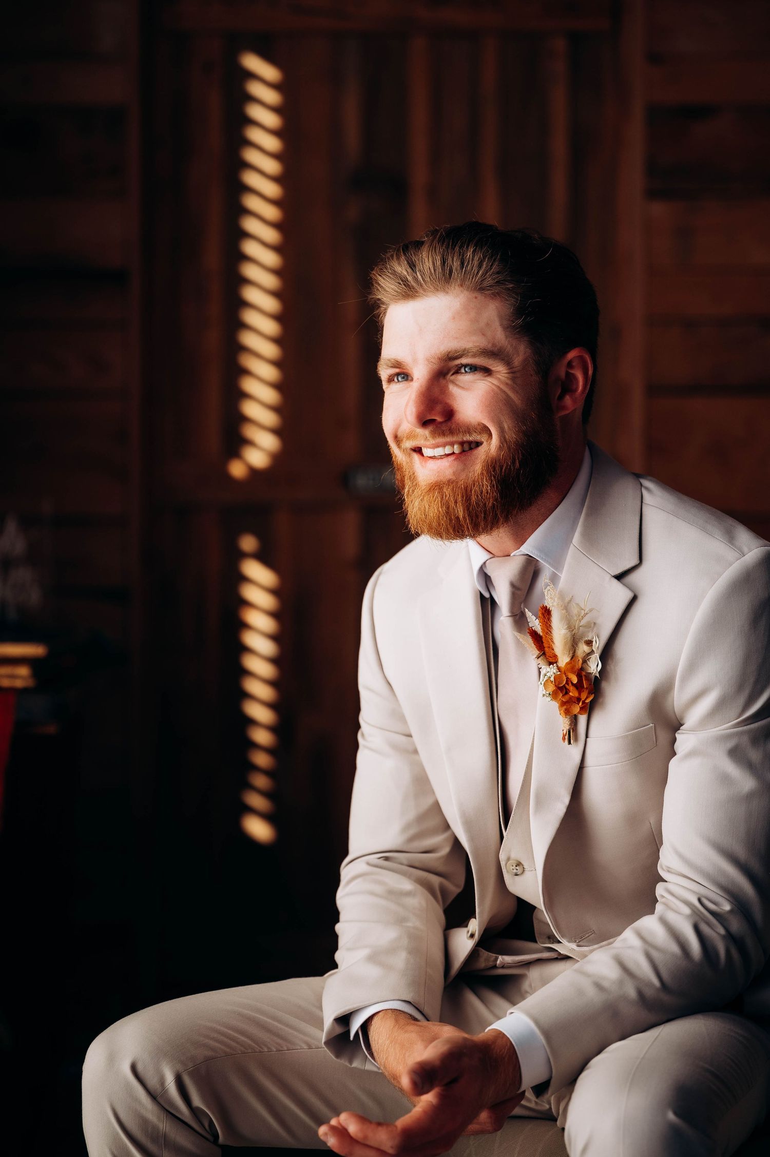 A groom in a cream colored suit sits indoors with string lights creating a warm ambiance.