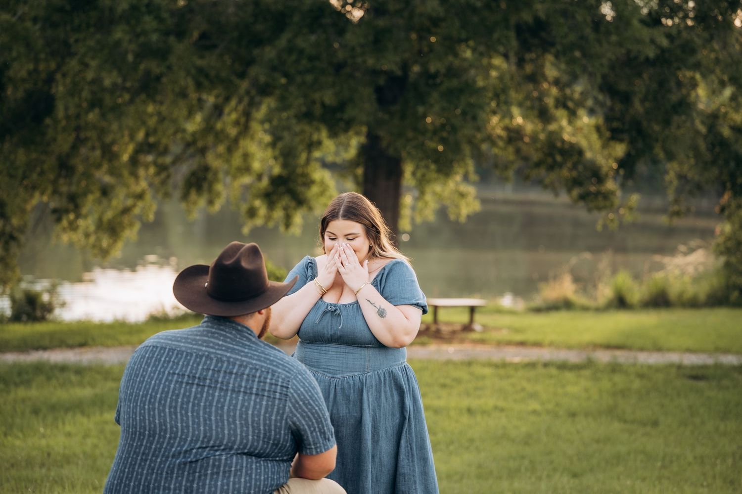 Man kneeling on grass proposing to surprised woman covering her mouth with hands near a pond.
