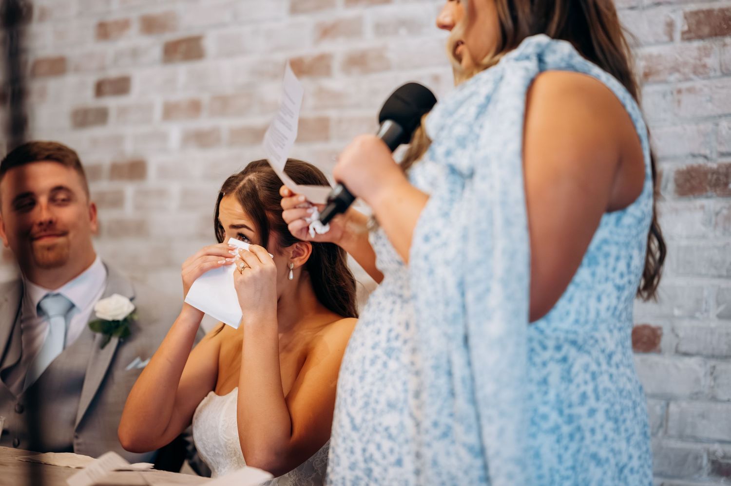 Bride wipes tears while maid of honor gives an emotional wedding reception speech at elegant venue.