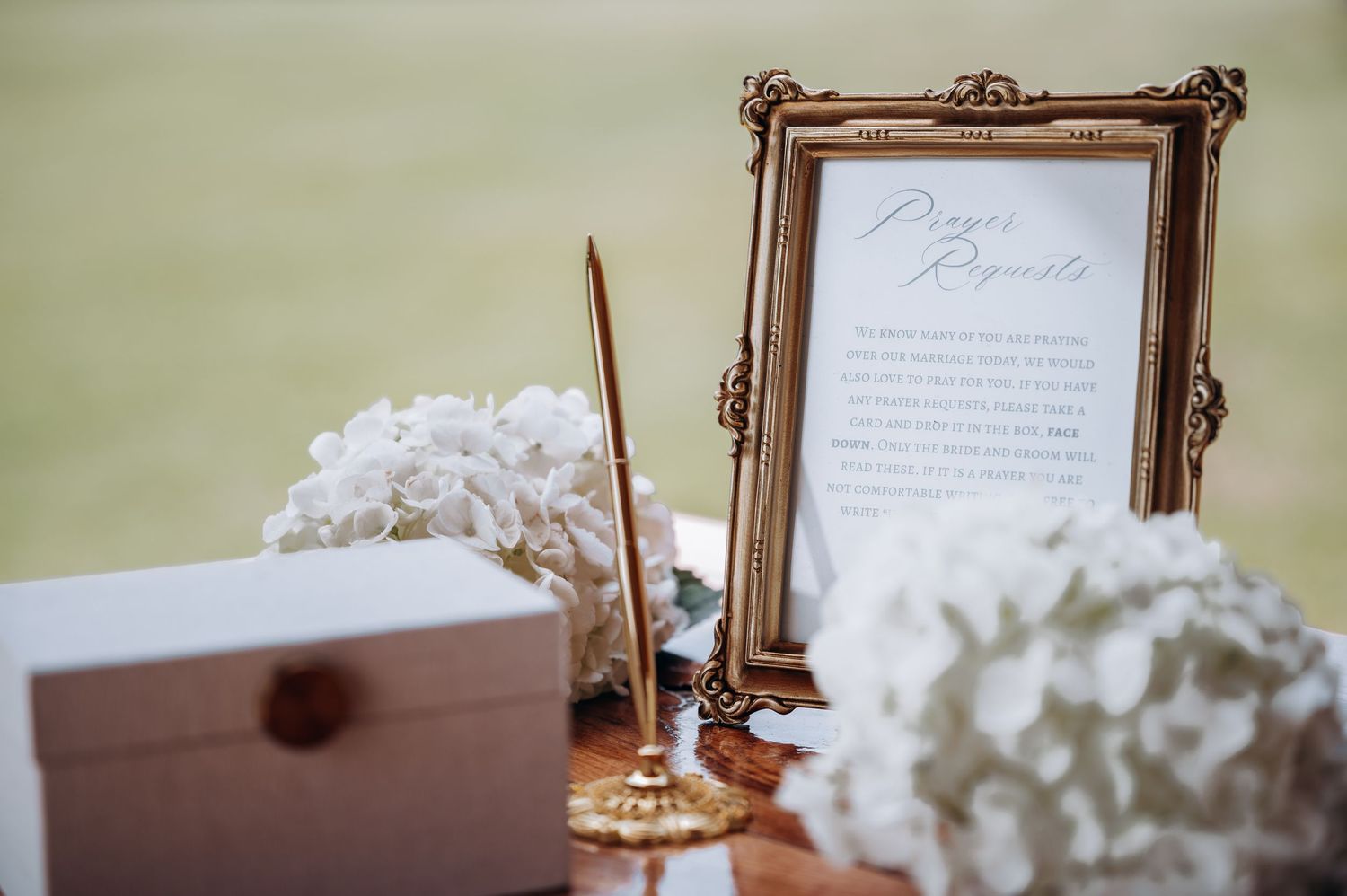 Elegant wedding prayer request sign in gold frame with white hydrangeas and a decorative pen on a table.