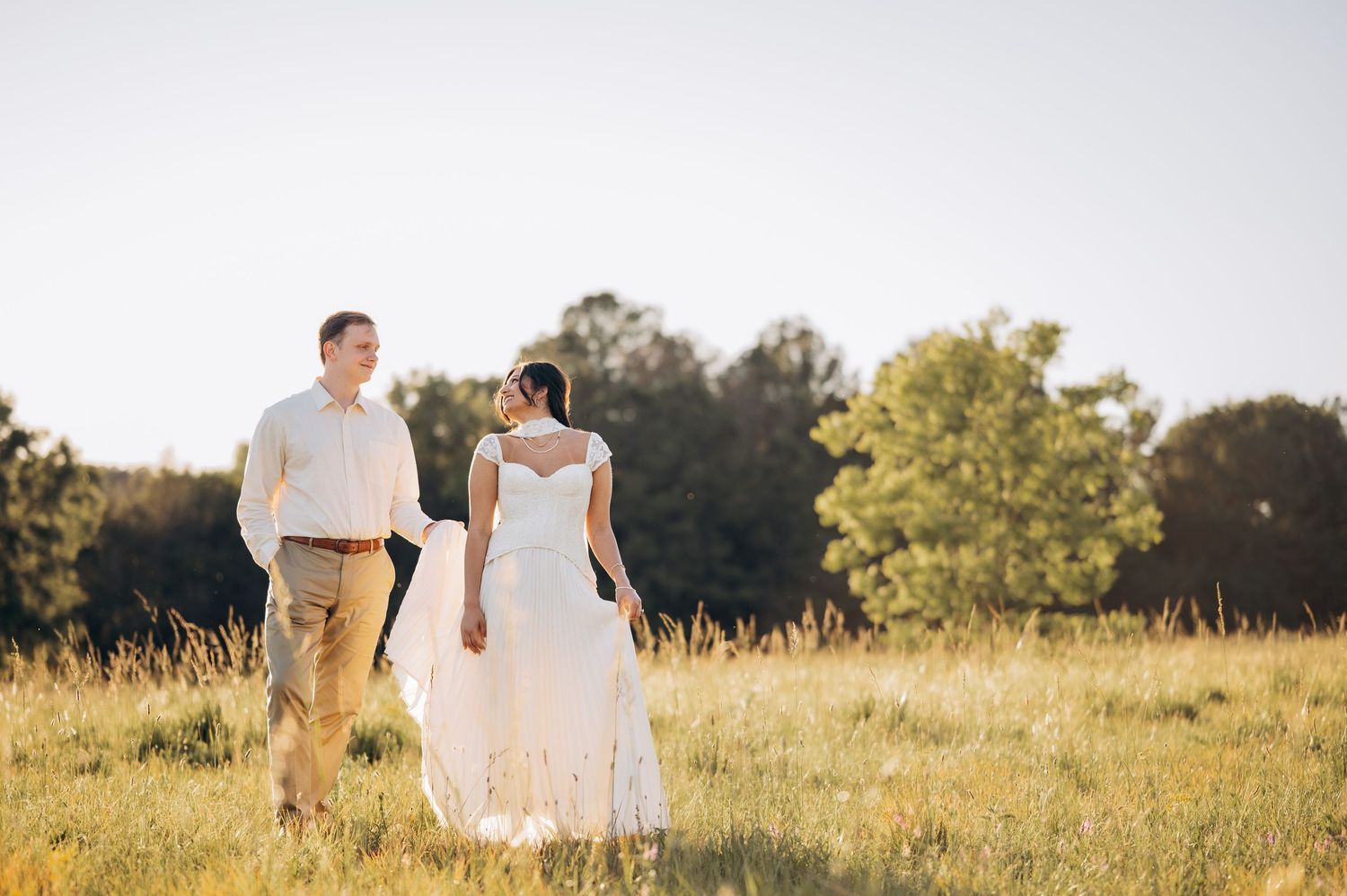 Couple strolling through a golden meadow on their wedding day, bride in white dress, sunny outdoor setting.