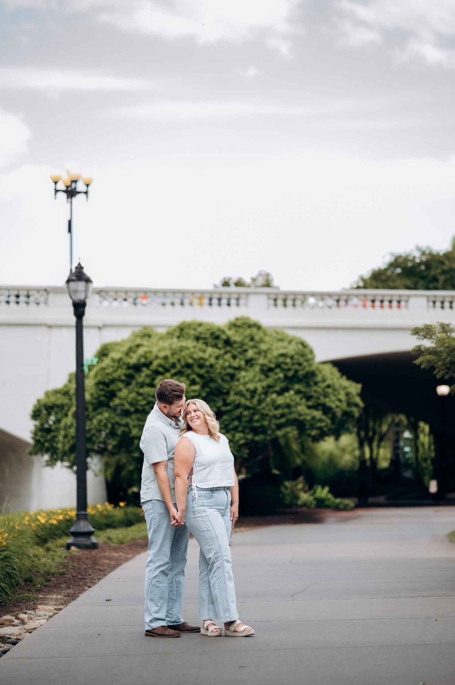 Couple in jeans sharing a romantic moment on a scenic pathway with a lamppost and bridge in the background.