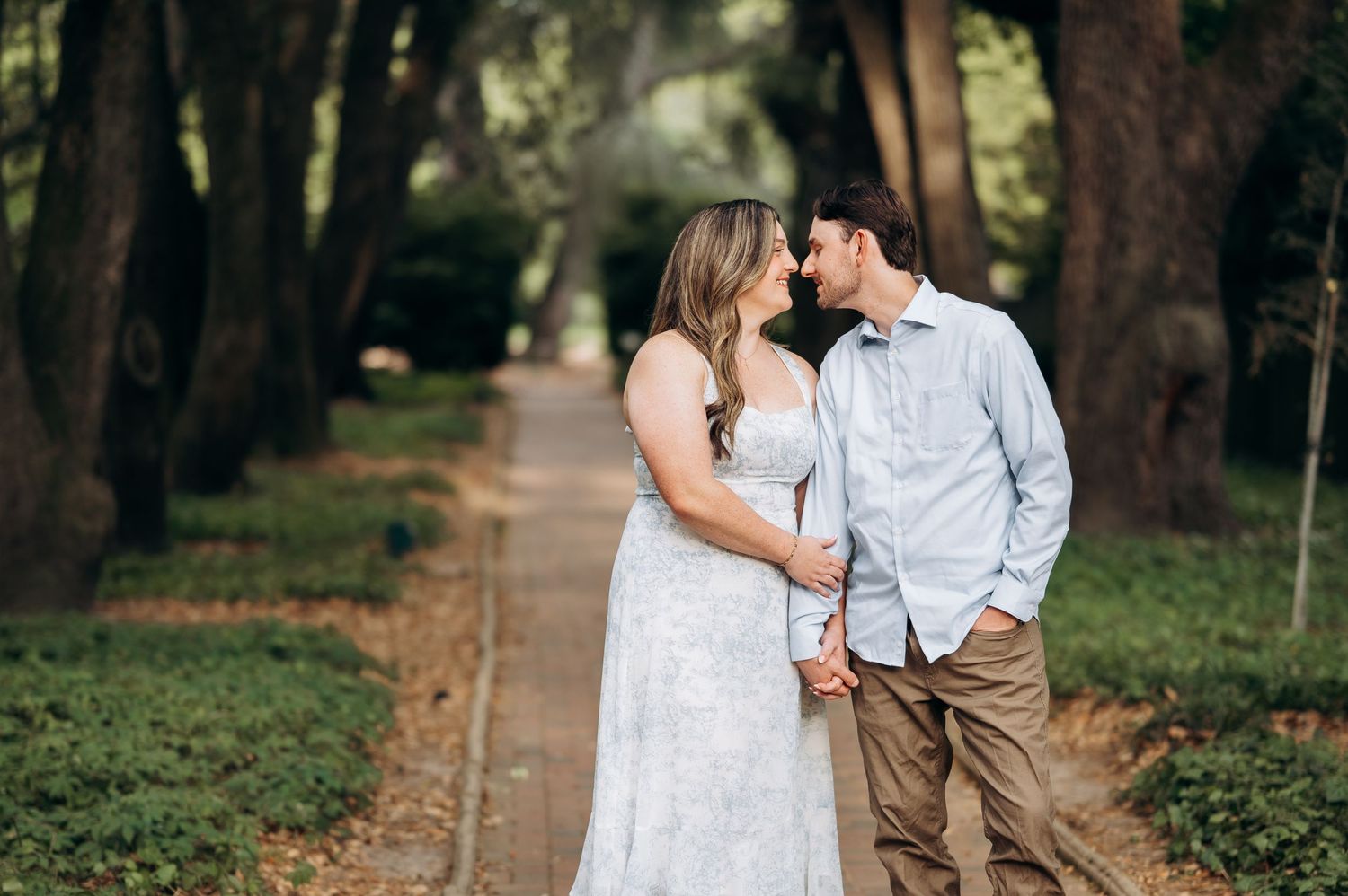 Couple sharing a romantic moment on a tree-lined path, woman in white dress and man in blue shirt holding hands.