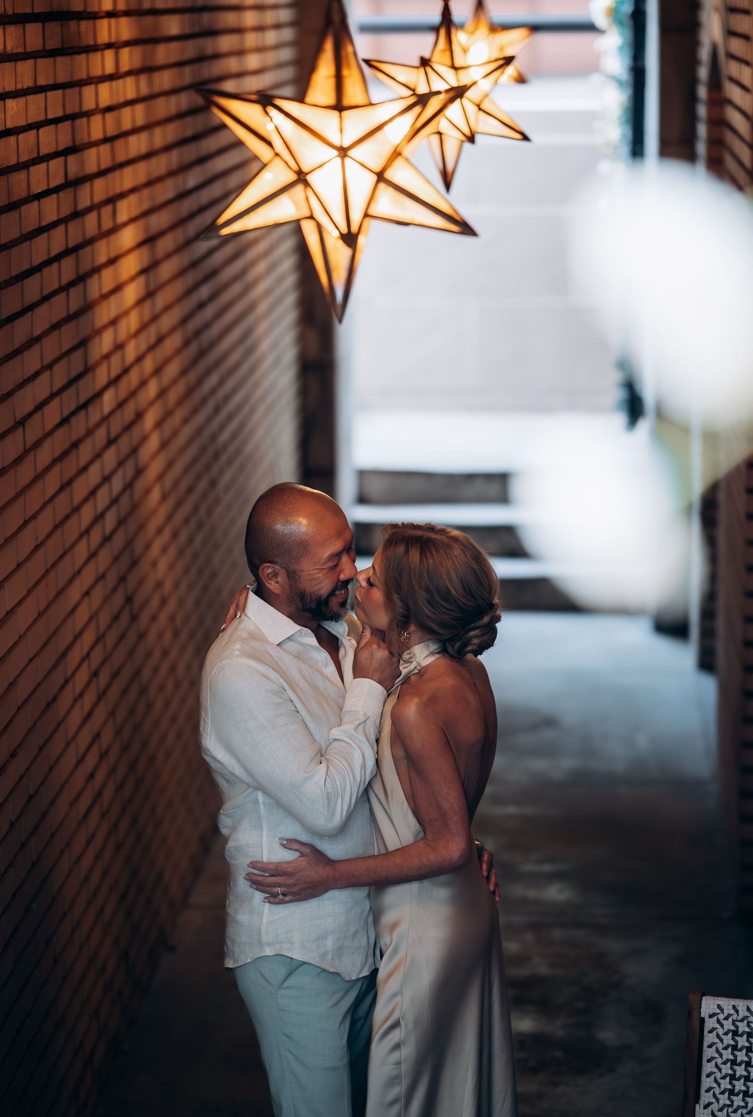 Couple sharing an intimate moment in a brick alley under glowing star-shaped pendant lights at night.