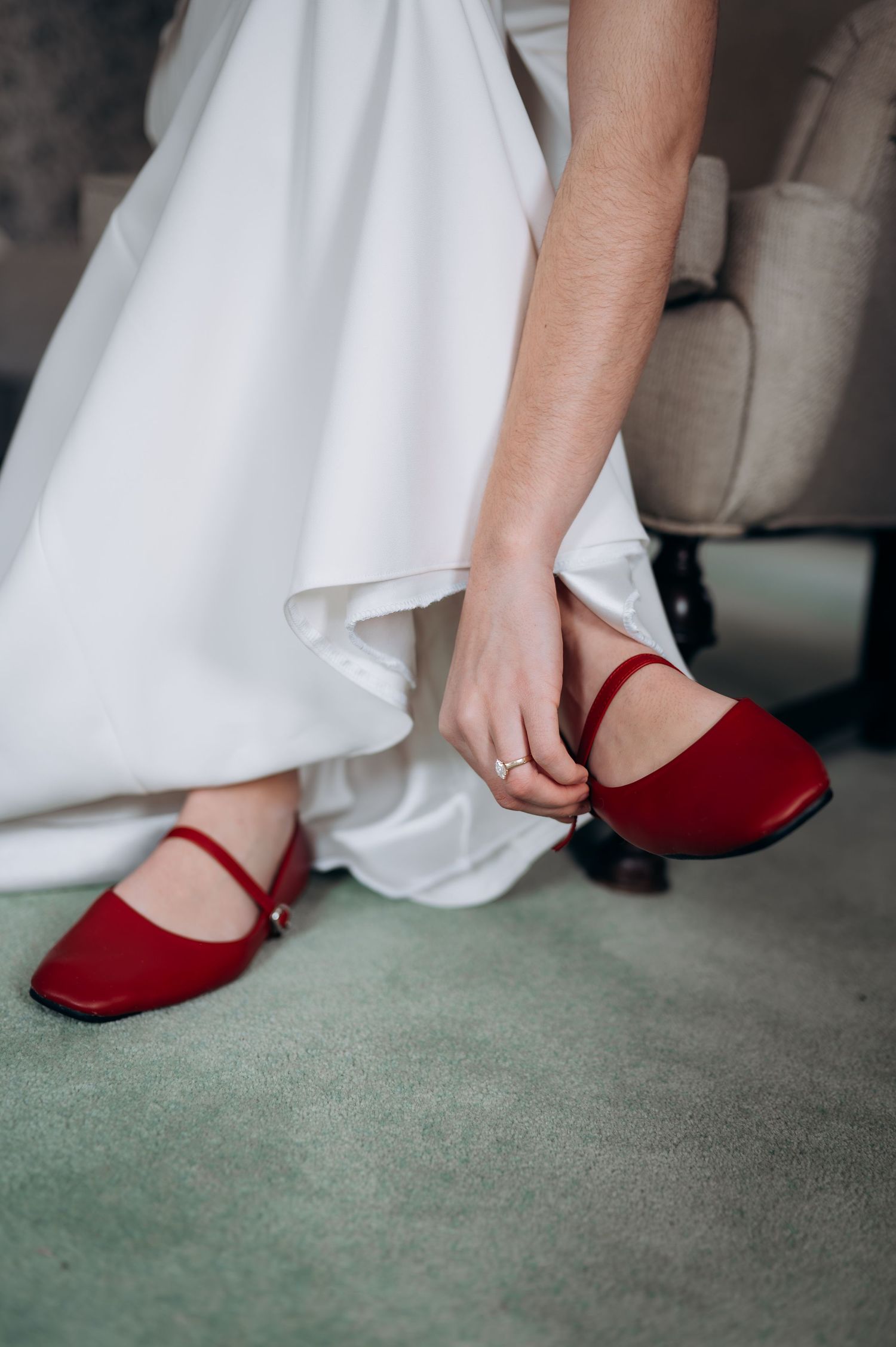 Bride adjusting red Mary Jane wedding shoes beneath white bridal gown while seated.