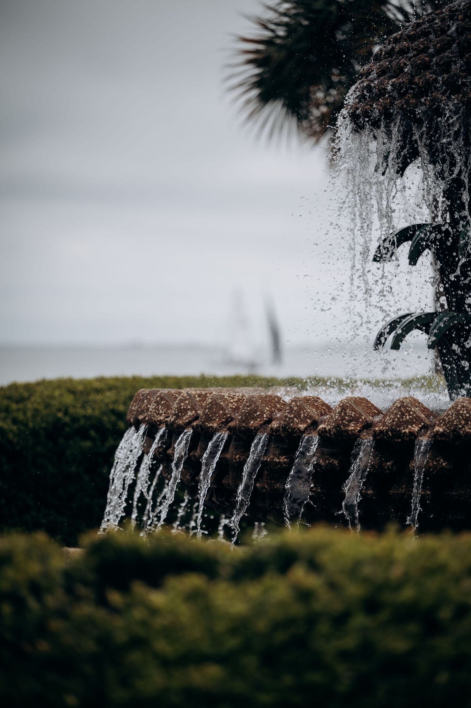 Water cascading over tiered stone fountain with palm tree and ocean in soft-focus background.