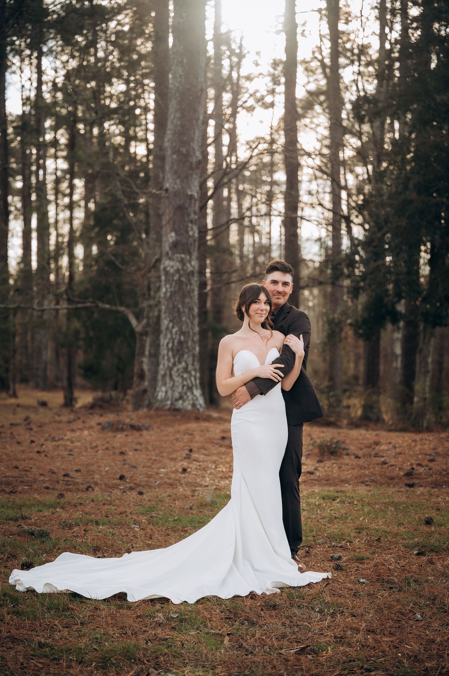 Wedding couple poses in forest setting with long white dress trailing on pine-covered ground.