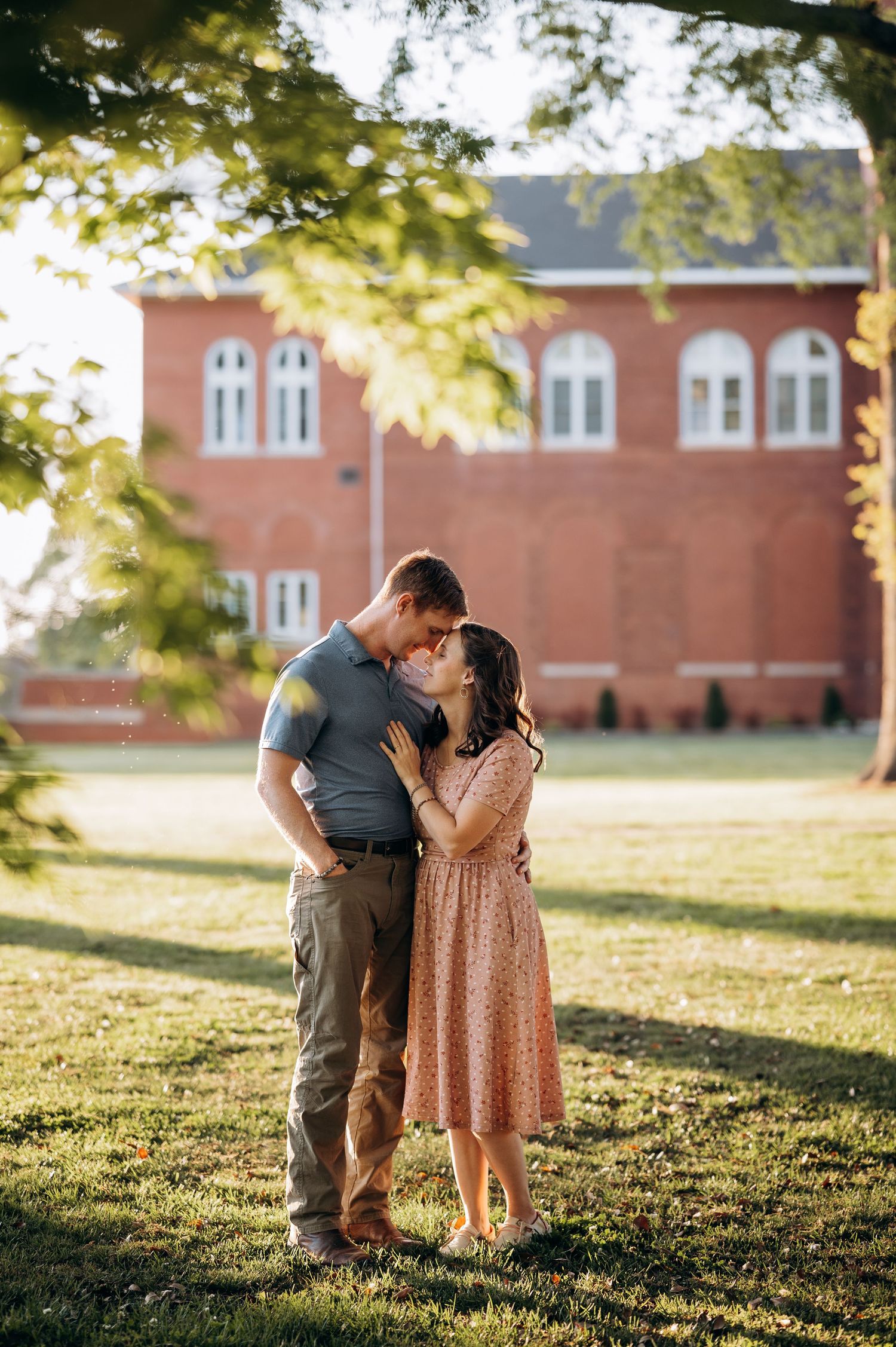 Couple sharing a romantic moment on green grass with a red brick building in the background on a sunny day.