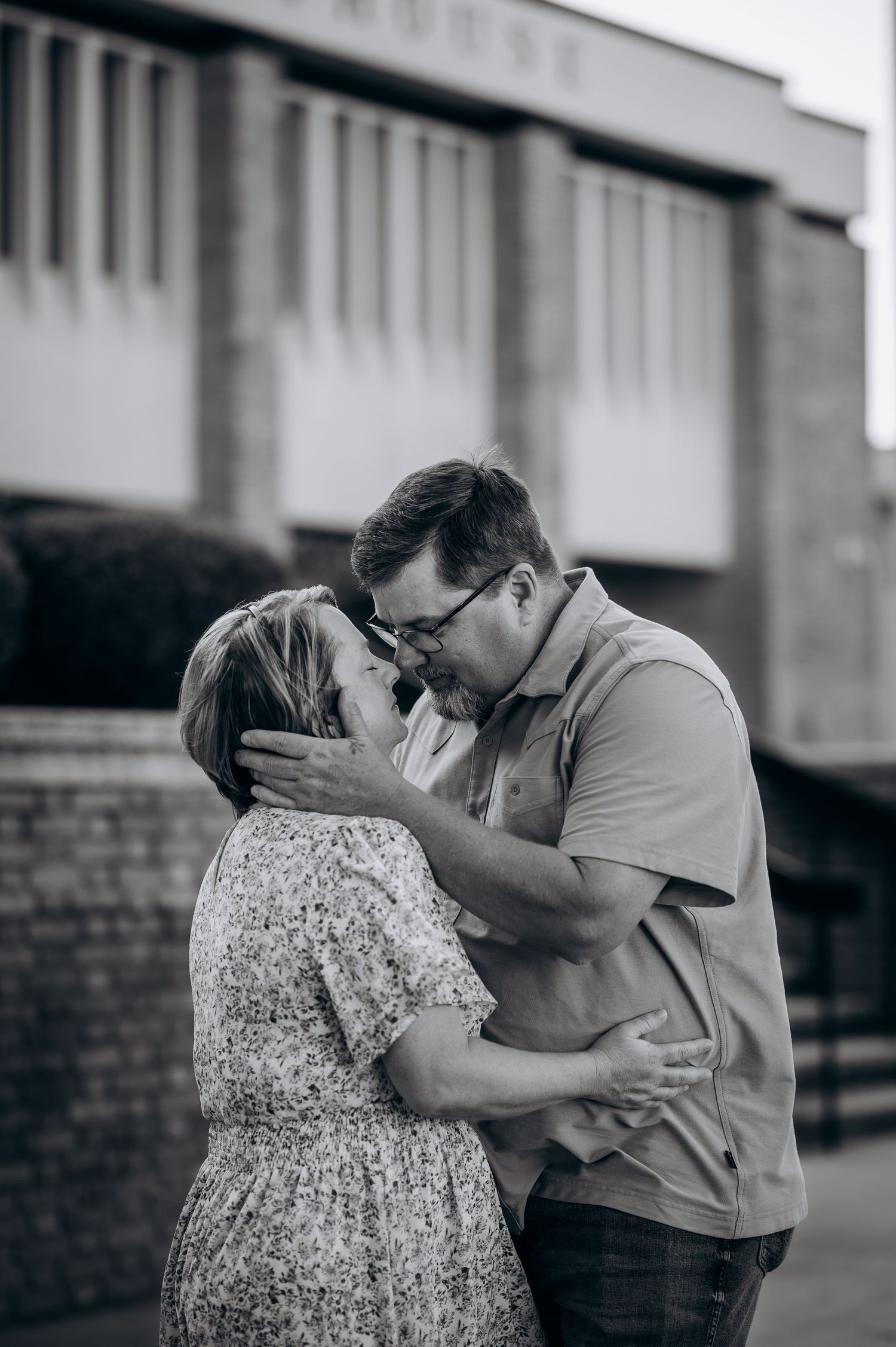 Black and white photo of a couple sharing a tender kiss outside a brick building.