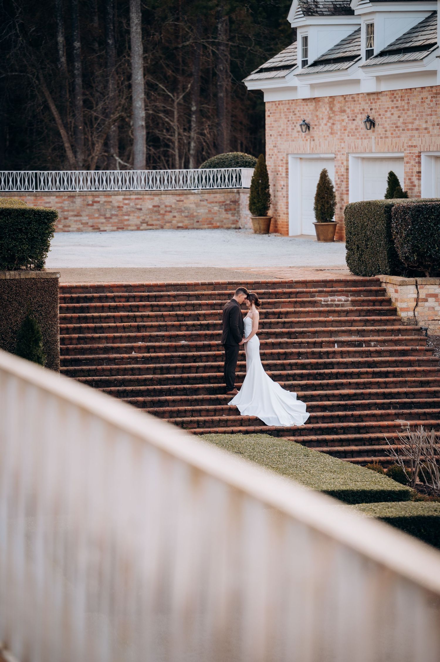 Bride and groom embrace on brick steps of elegant estate venue, surrounded by manicured hedges and topiaries.