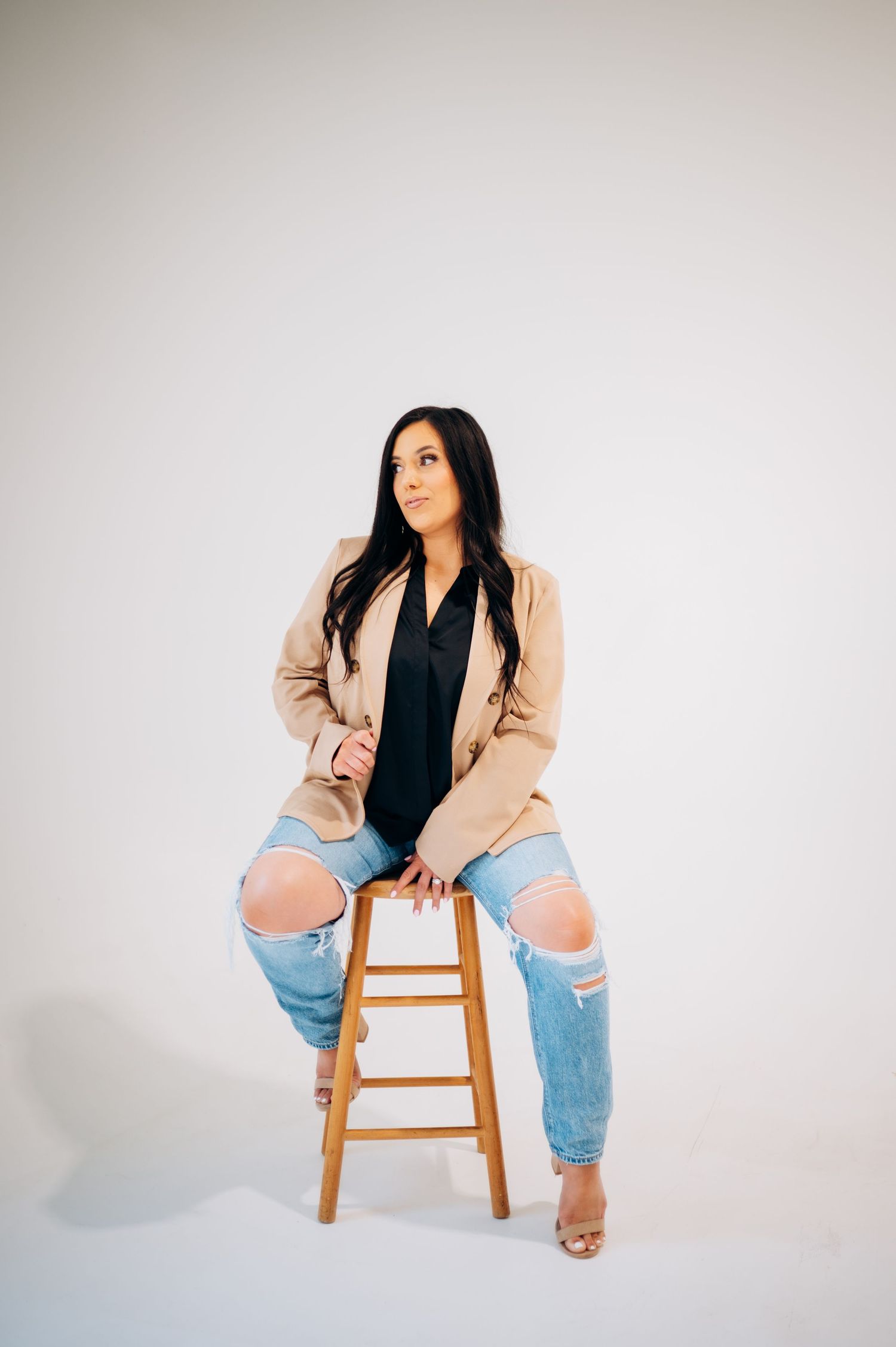 A person in a beige blazer and distressed jeans sits casually on a wooden stool against a white backdrop.