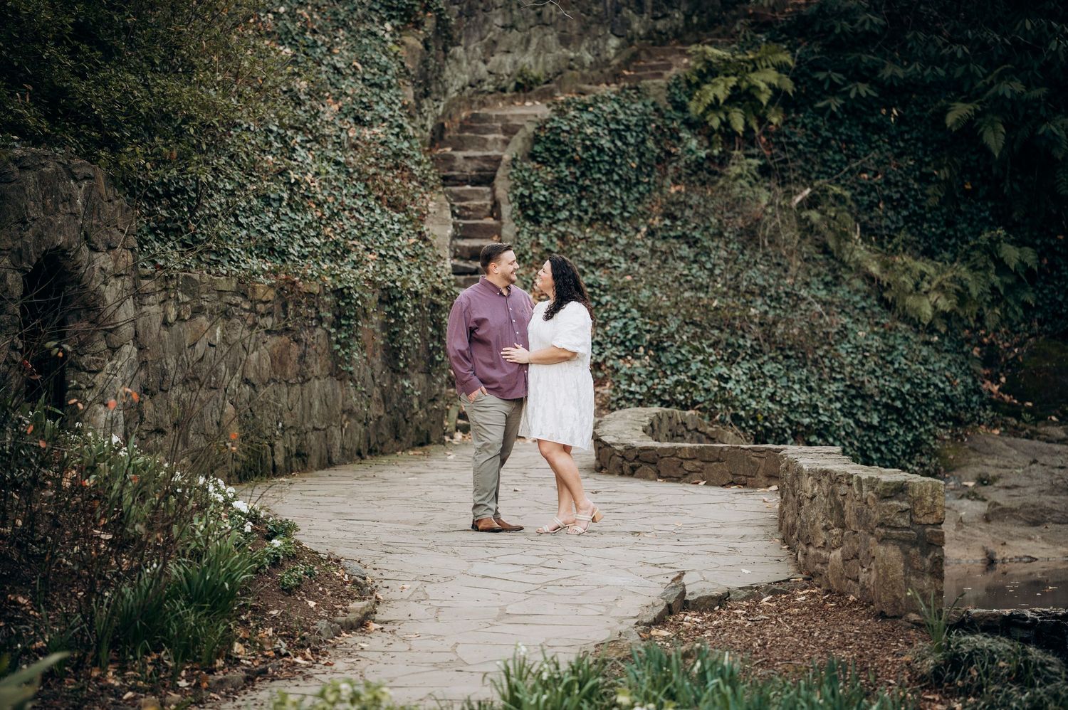 A couple sits together on stone steps in a garden path surrounded by lush greenery and rustic stone walls.