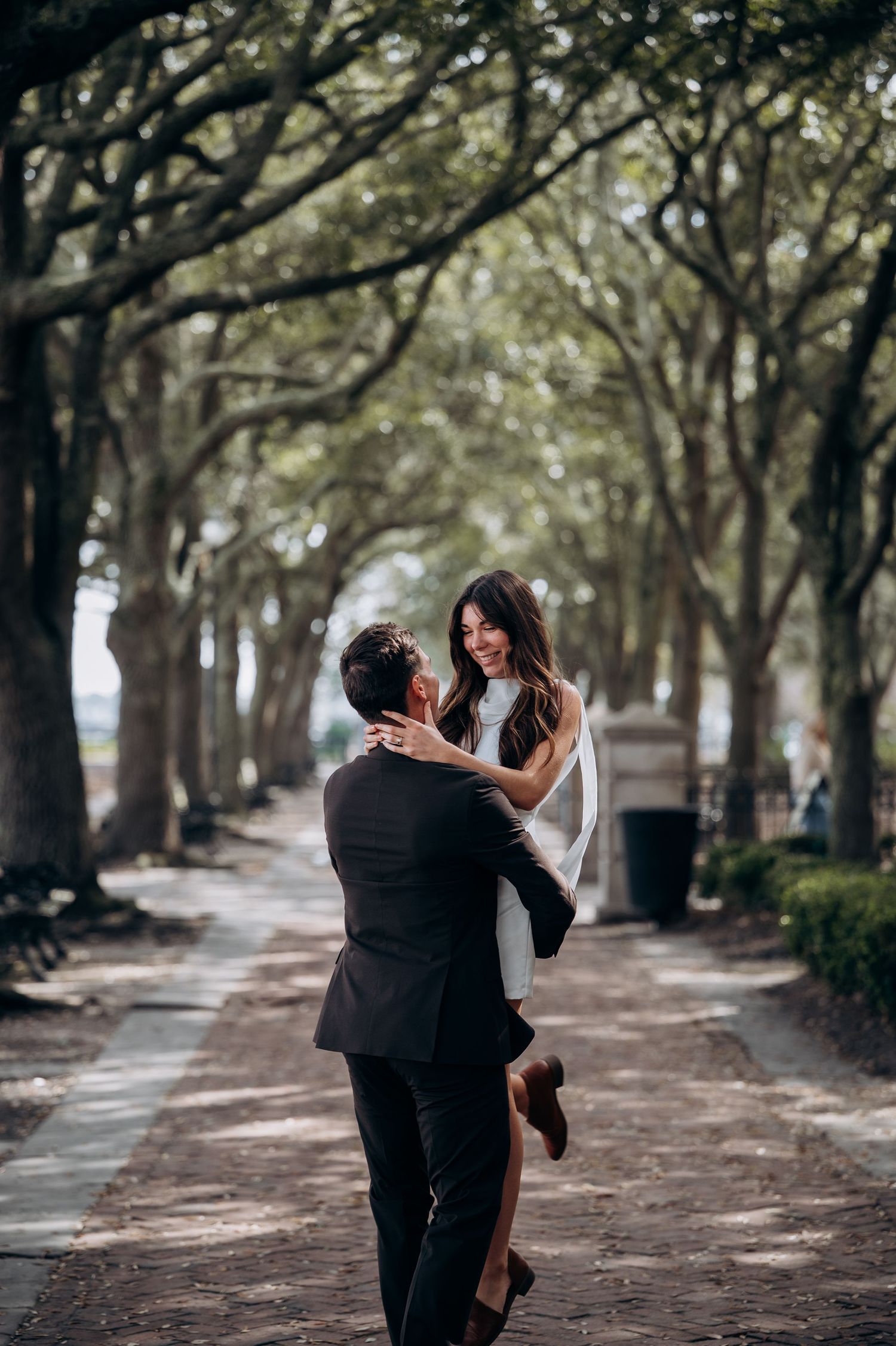 A couple dances playfully along a tree-lined pathway on a sunny day.