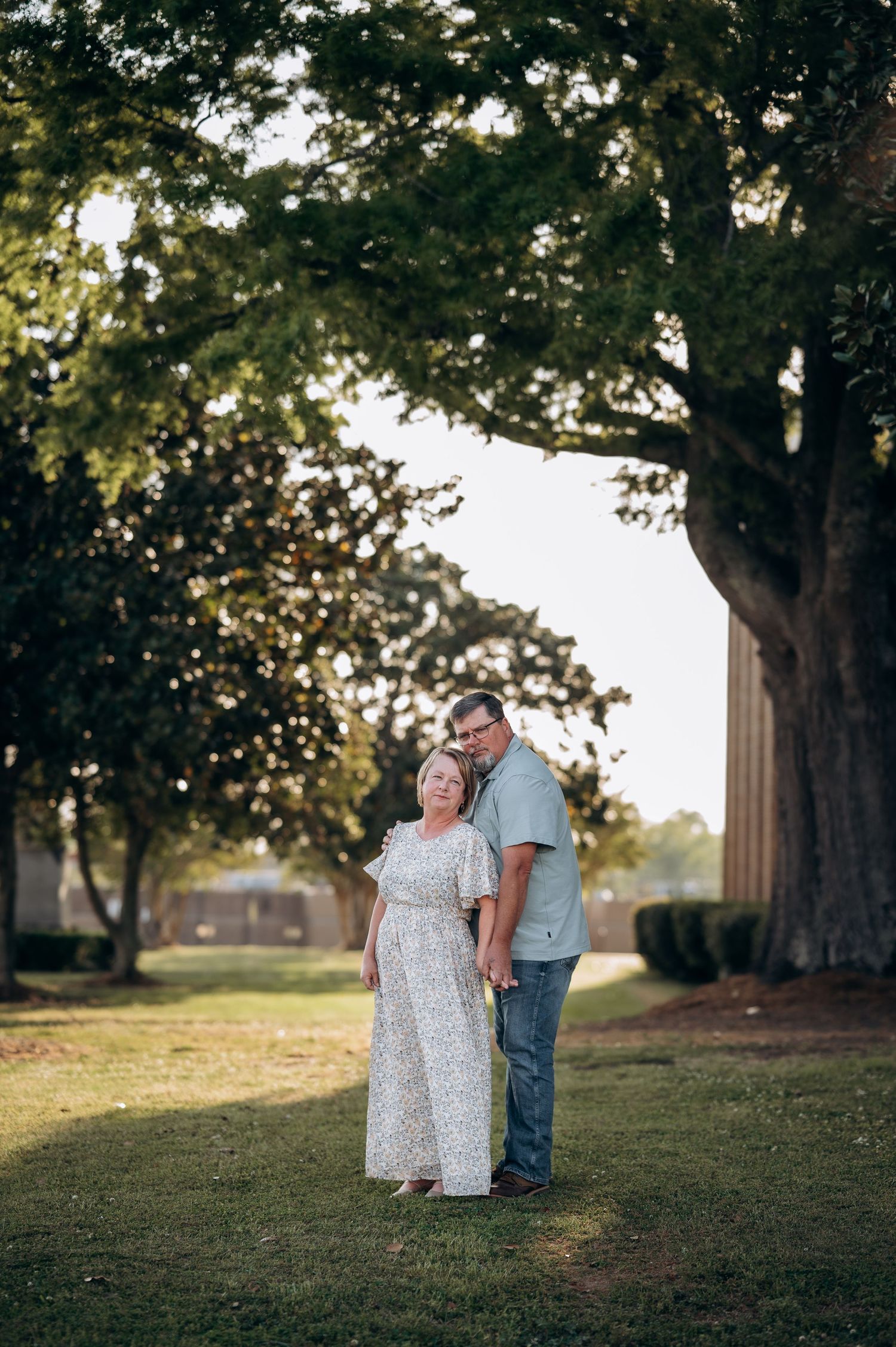 Couple posing together under large oak trees on green lawn during golden hour outdoor portrait session.