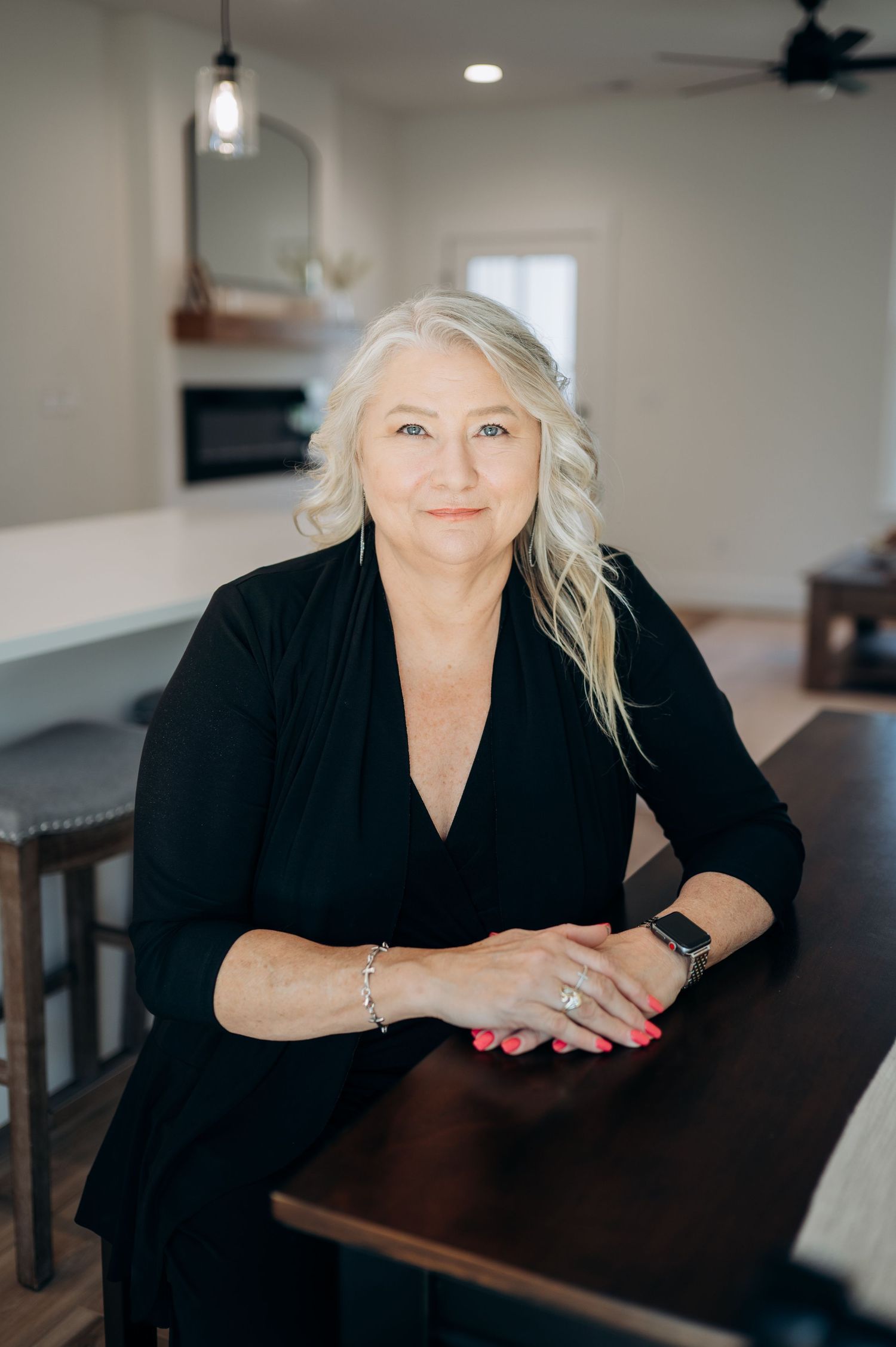 A smiling middle-aged woman with blonde hair wearing a black outfit leans on a wooden table in a modern home interior.