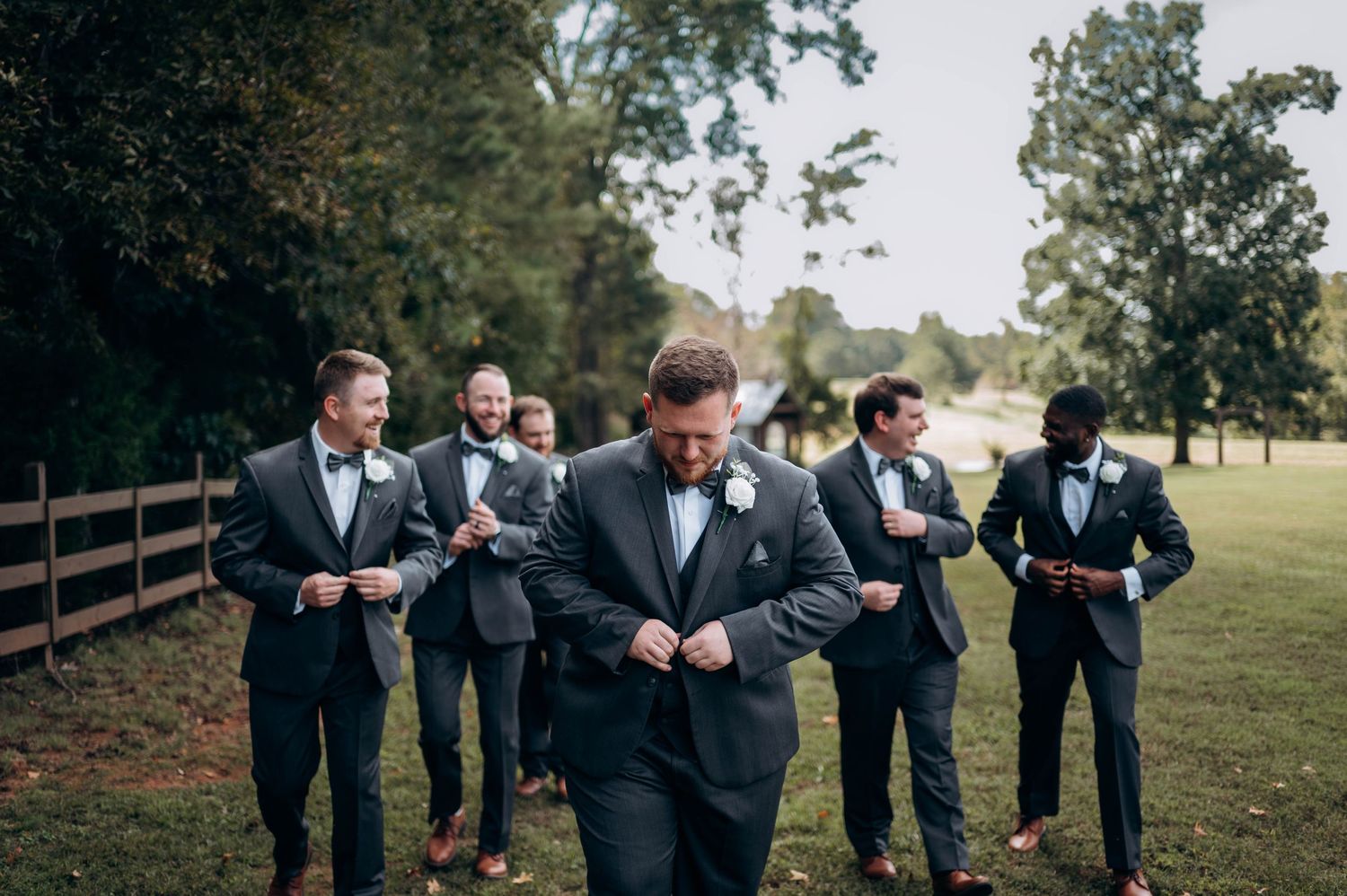 Wedding party in black tuxedos poses playfully on grassy outdoor setting.