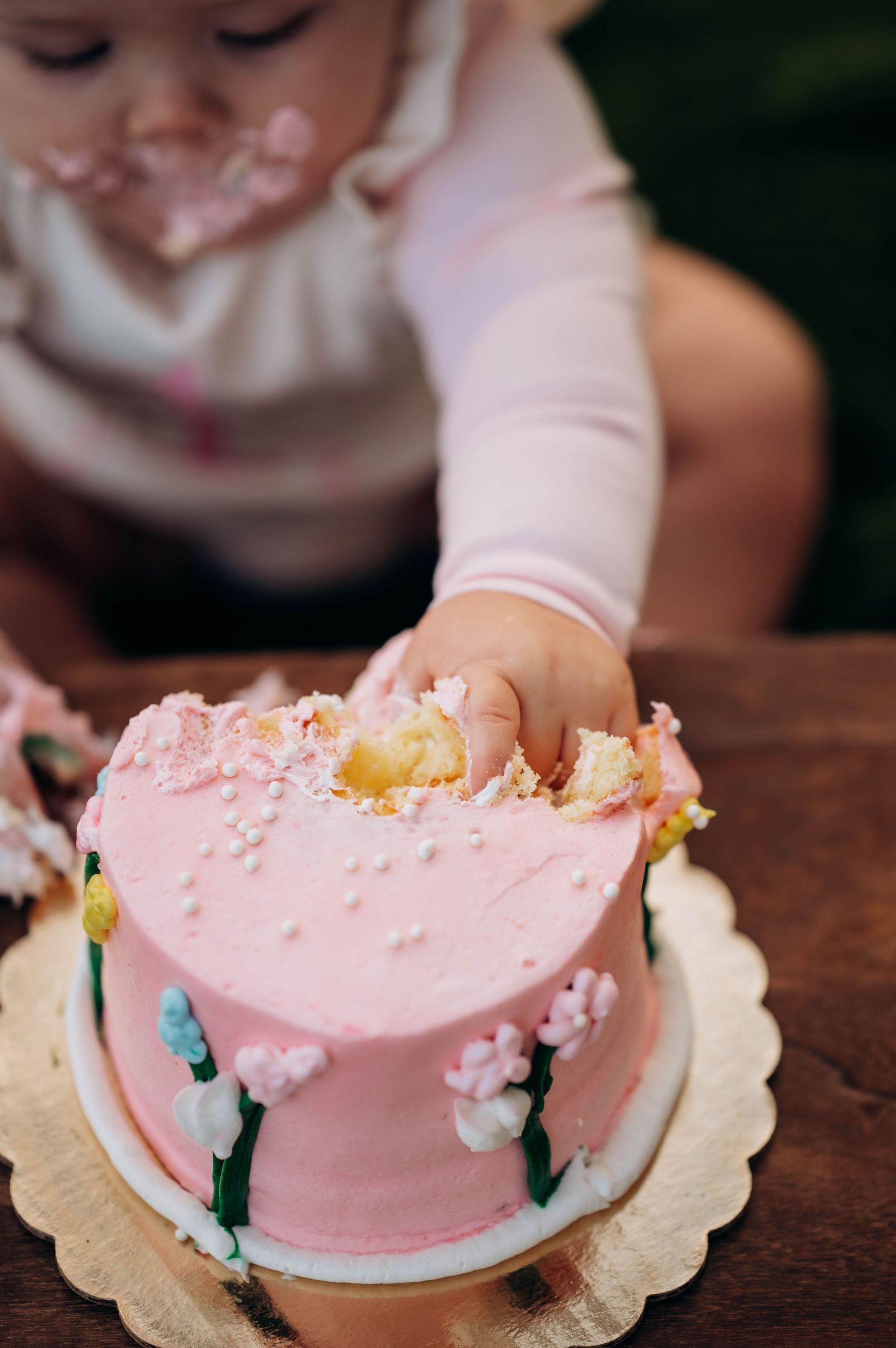 Small hands reach for a partially eaten pink birthday cake on a wooden surface.